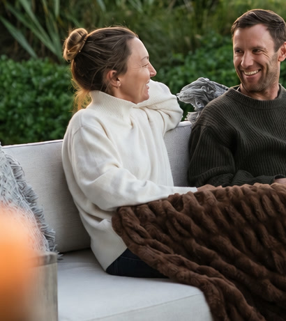 Ana Lynch, General Manager of Bluestone Estate, in a cream jumper on the deck sofa at dusk with a glass of red wine, laughing with her husband Ben under a brown throw, flax and hedges behind at sunset.