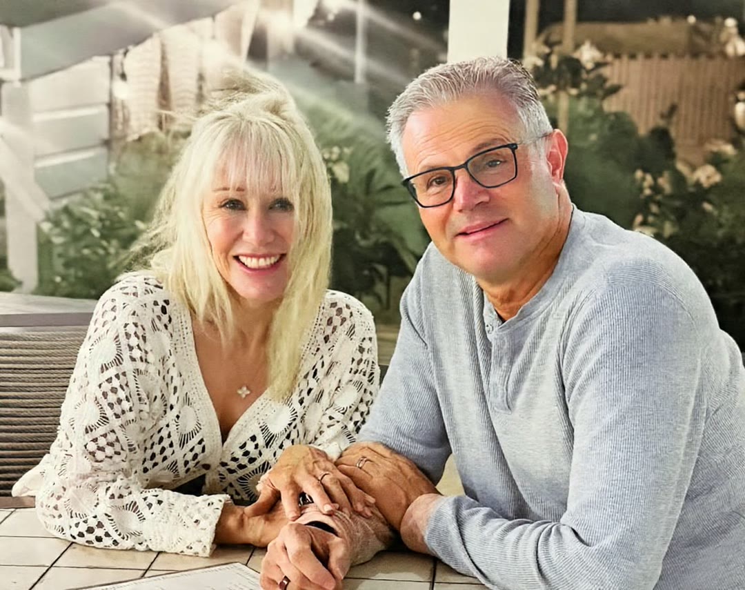 Andrea Grant Robbiati and Tarek Robbiati, owners of Bluestone Estate, side by side at a table on a terrace in the evening, Andrea in a white crochet top and Tarek in glasses and a grey top, hands together.