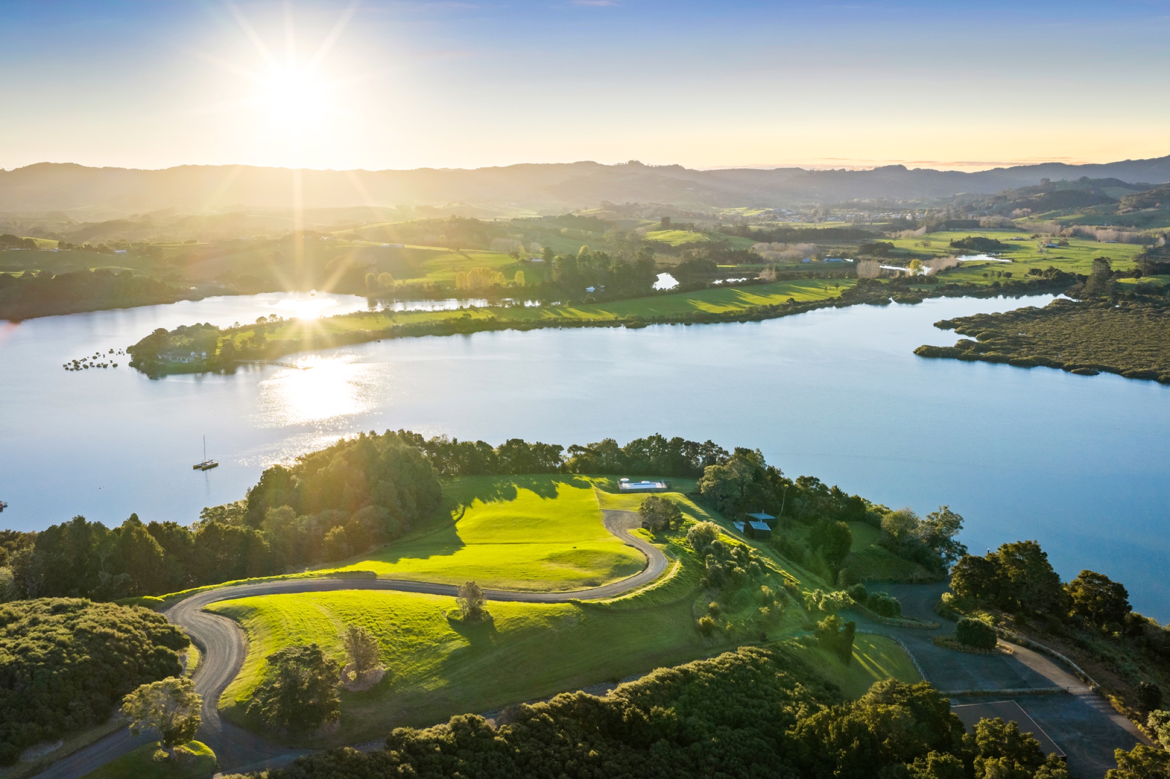 Bluestone Encampment from the air at sunset: the headland's lawn and drive above the inlet, the pool pavilion at the edge of the bush, a yacht at anchor, and the Matakana hills across the water.