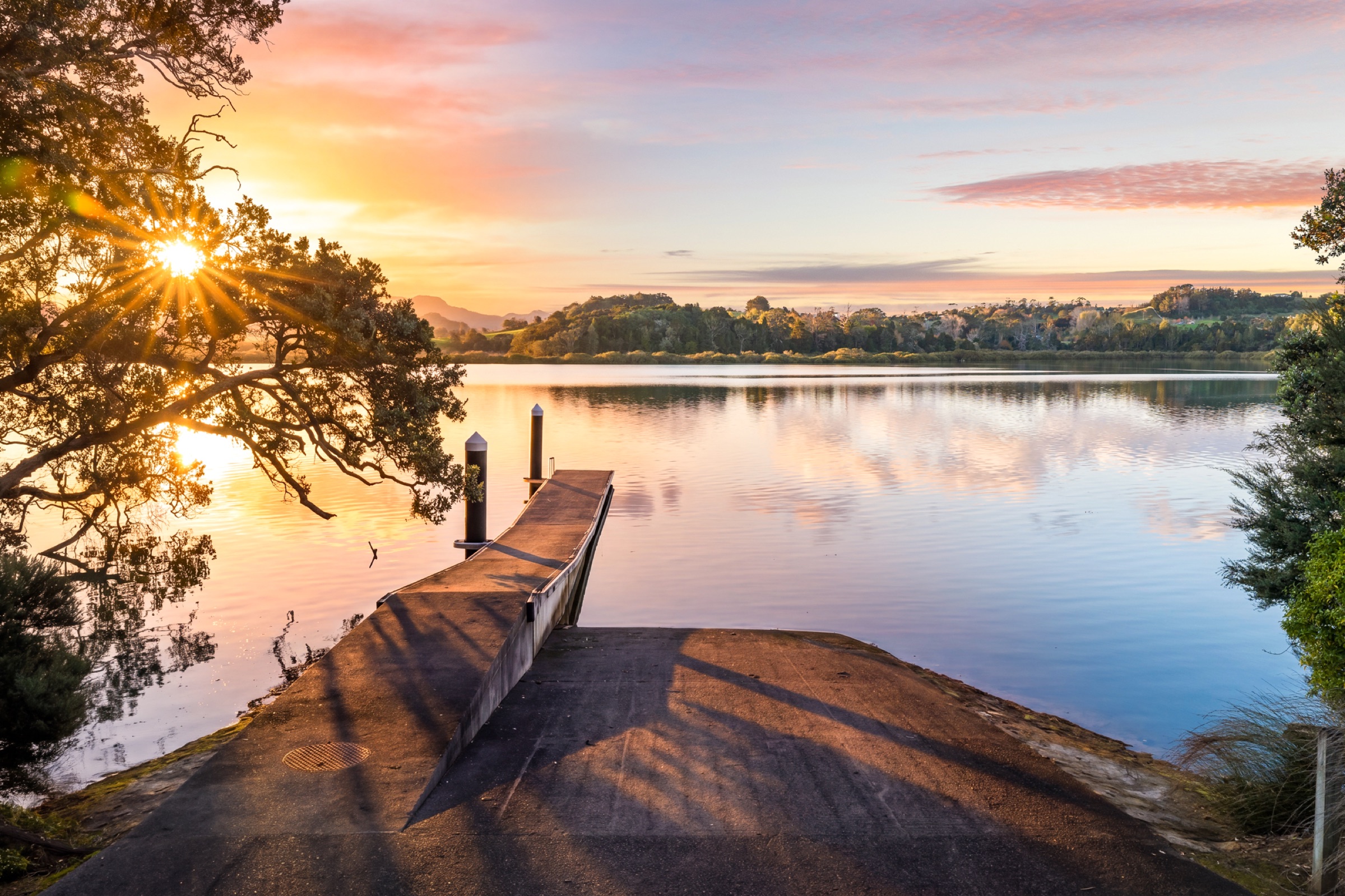 The private boat ramp and jetty at Bluestone Encampment at sunrise, the inlet flat calm, the sun through a pōhutukawa, and the far shore's bush and farmland reflected in the water.