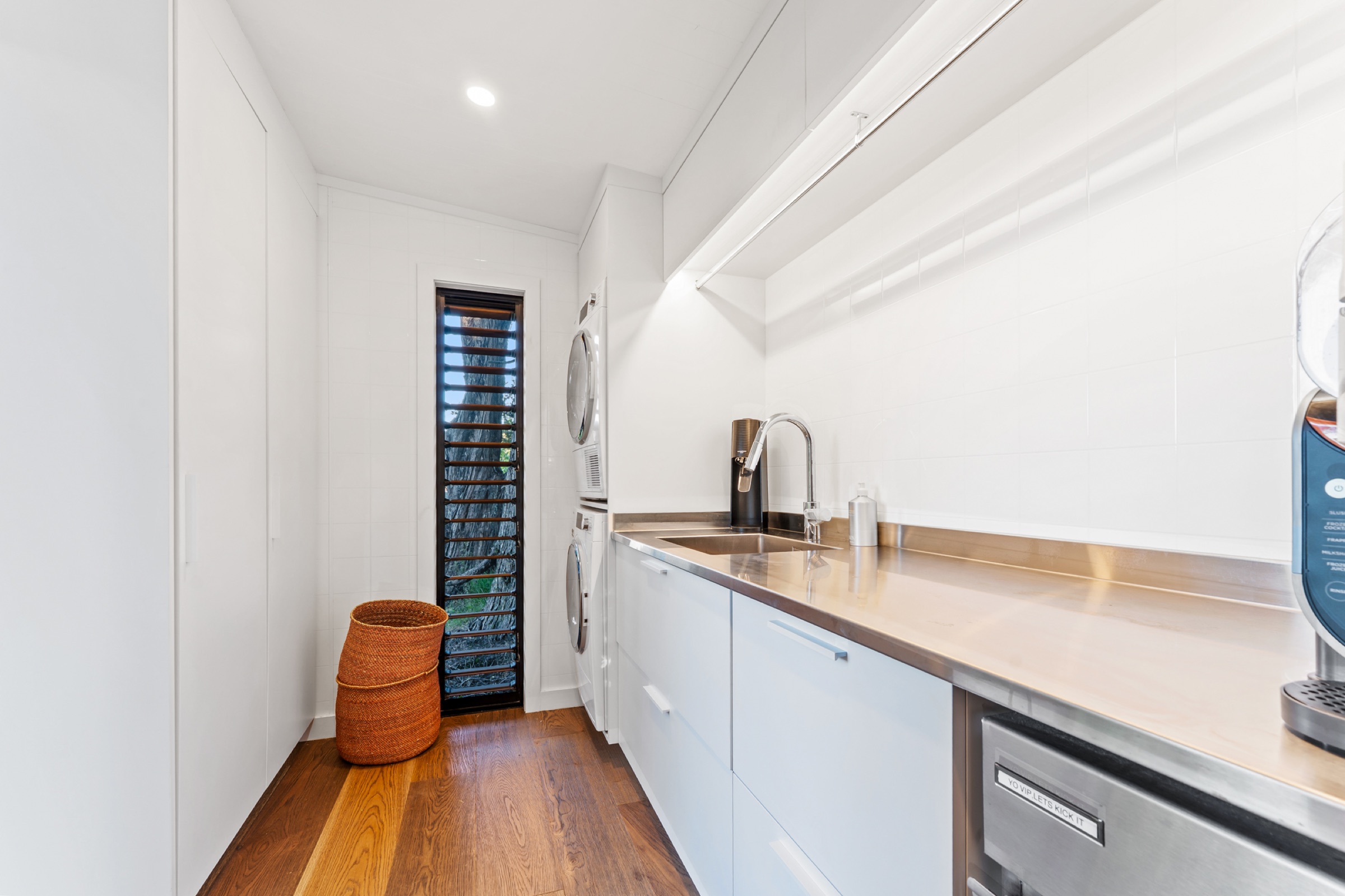 The laundry pavilion at Bluestone Encampment: a stacked washer and dryer, a stainless bench with a sink, white cabinetry, and a louvre window onto the bush.