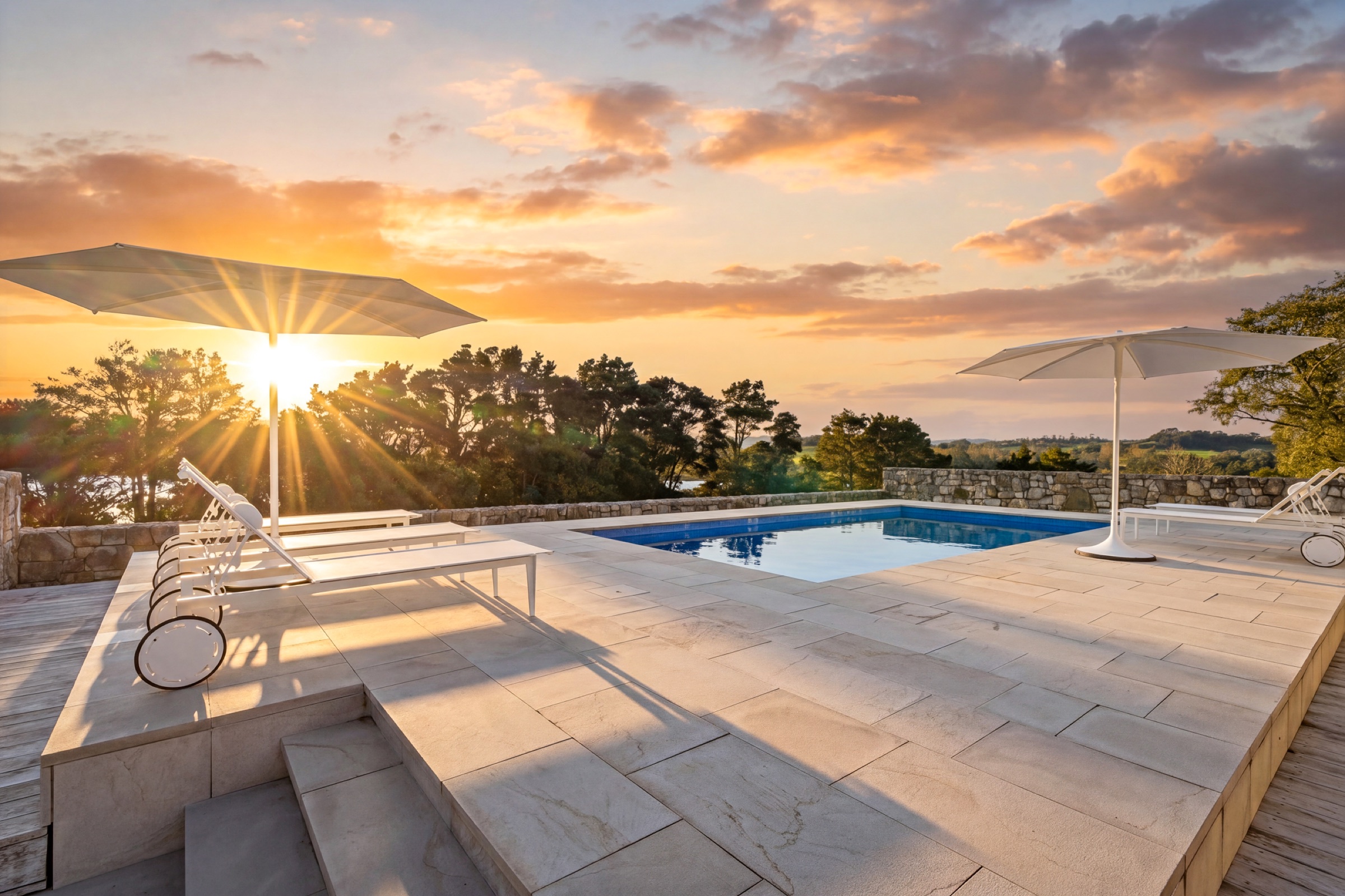 The pool at Bluestone Encampment at sunset: stone-paved terrace, white loungers and umbrellas, the stone wall, and the sun going down behind the trees over the inlet.