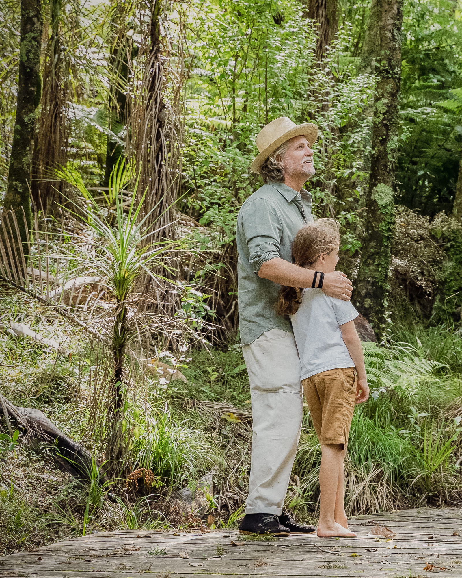 A guided bush walk at Bluestone Estate: a guide in a straw hat with a hand on a child's shoulder, both looking up into the canopy from a boardwalk among nīkau, tree ferns, and kānuka.