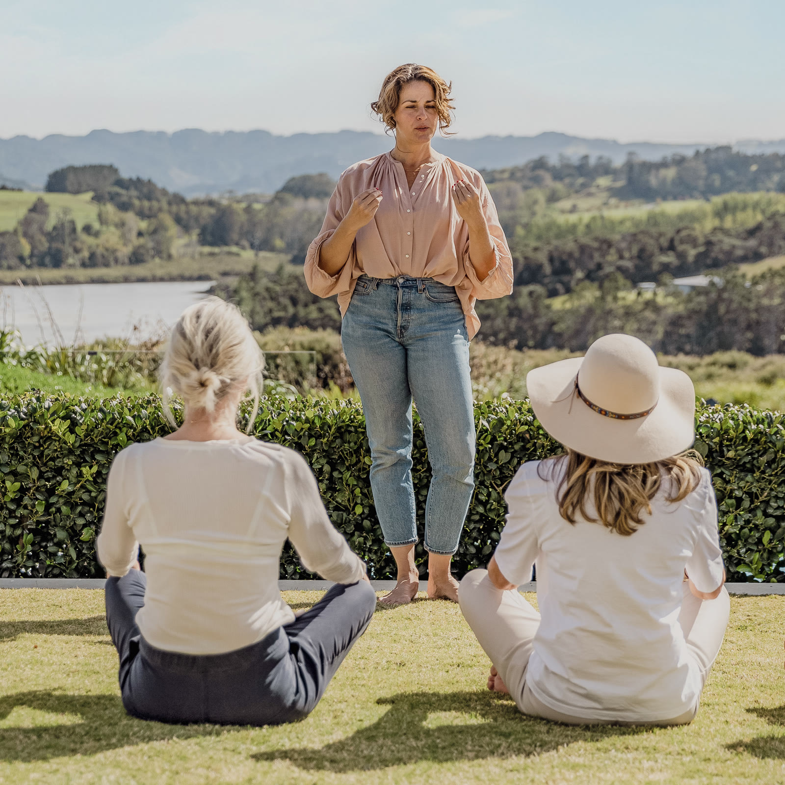 A guided breathwork and meditation practice on the lawn at Bluestone Estate: the teacher standing with eyes closed, two guests seated cross-legged before her, and the estuary and hills beyond.