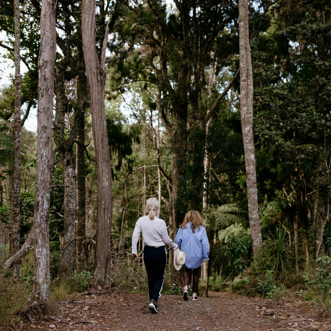 Two guests walking away down a gravel bush track at Bluestone Estate under tall native trees, one carrying a straw hat, tree ferns along the path.