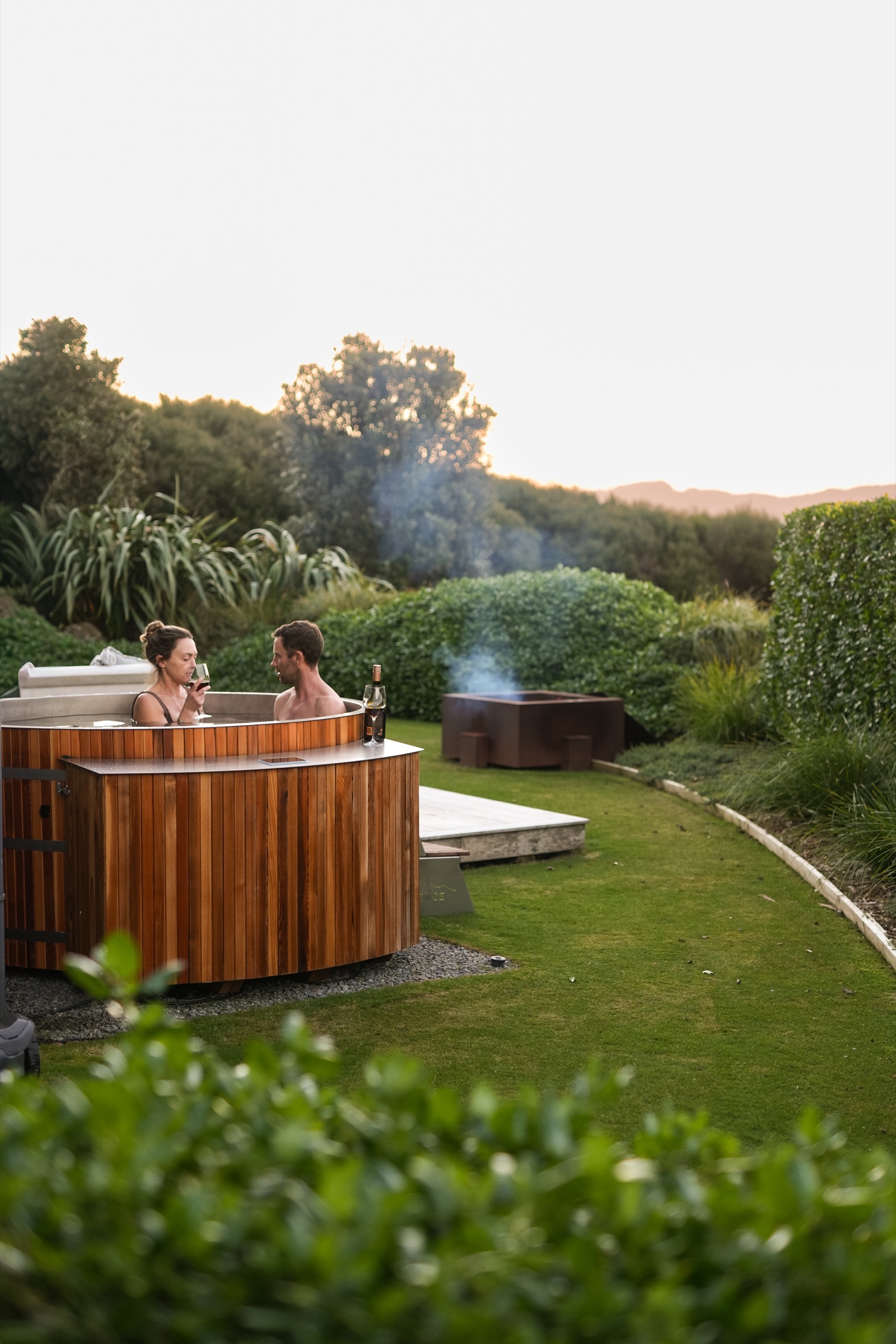 A couple with a glass of wine in the cedar hot tub on the Boat House lawn at Bluestone Estate at dusk, the steel firepit smoking behind them and the hedges and bush turning gold.