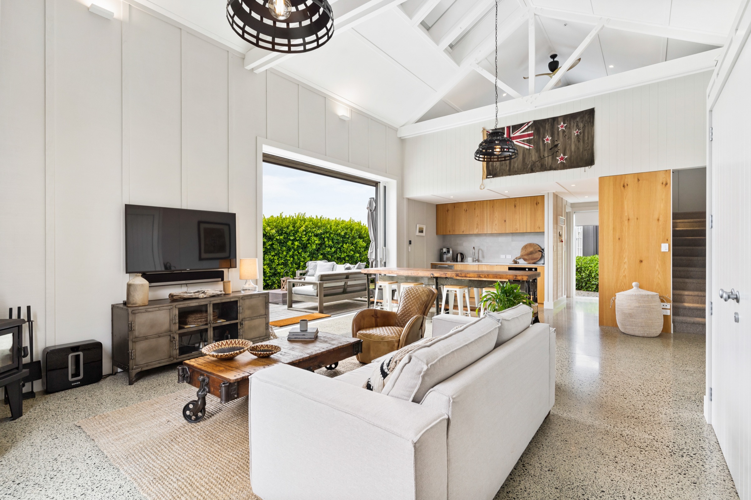 The open-plan living space of the Boat House at Bluestone Estate under its cathedral ceiling and skylights: a linen sofa, an industrial coffee table on castors, the oak kitchen with a long timber bar, and the doors open to the deck.