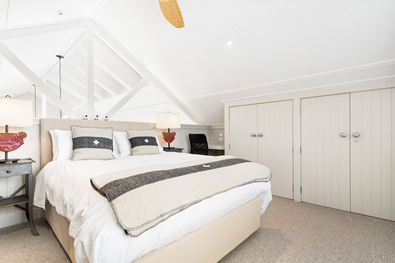 The loft bedroom of the Boat House at Bluestone Estate: a queen bed with Swiss cross cushions and a striped wool throw under white painted trusses, a ceiling fan, and pale panelled wardrobes.