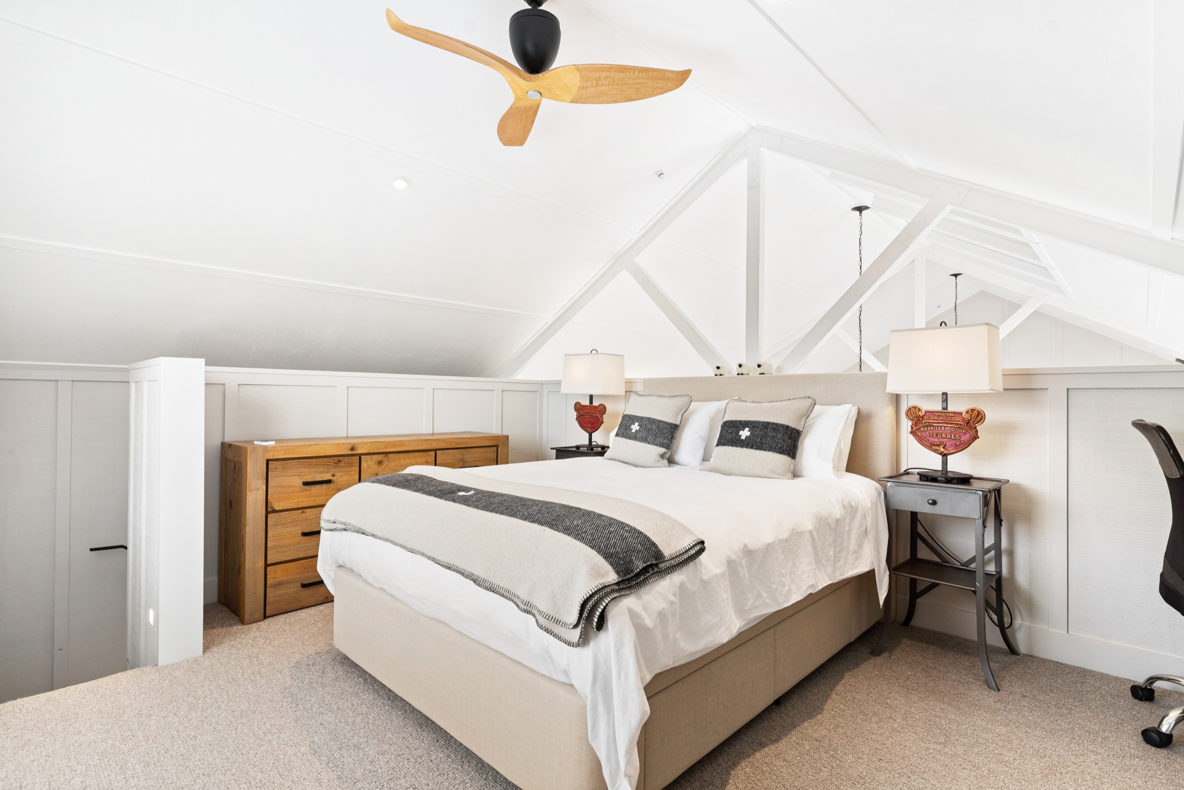 The loft bedroom of the Boat House at Bluestone Estate: a queen bed with a grey wool blanket under the white timber trusses, a ceiling fan, and a timber chest of drawers.