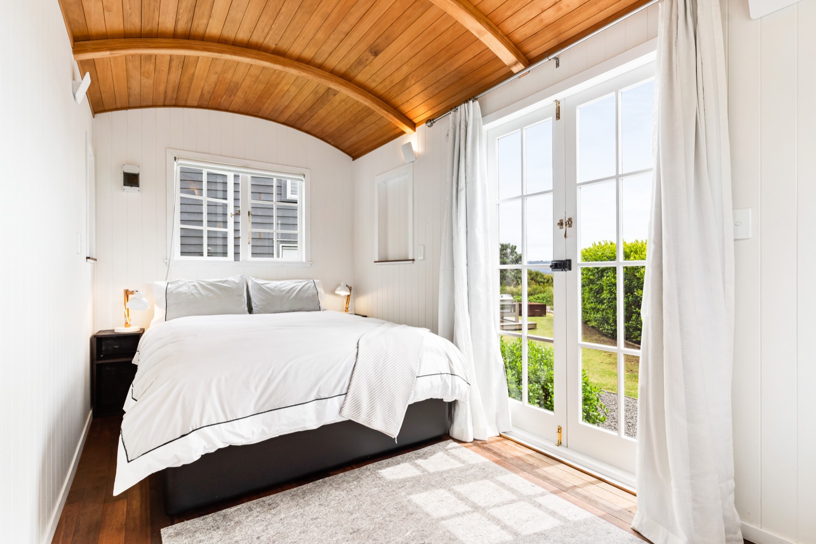 The Shepherd's Hut at Bluestone Estate: a queen bed under a curved kauri ceiling, white panelled walls, and French doors open to the lawn and the firepit.