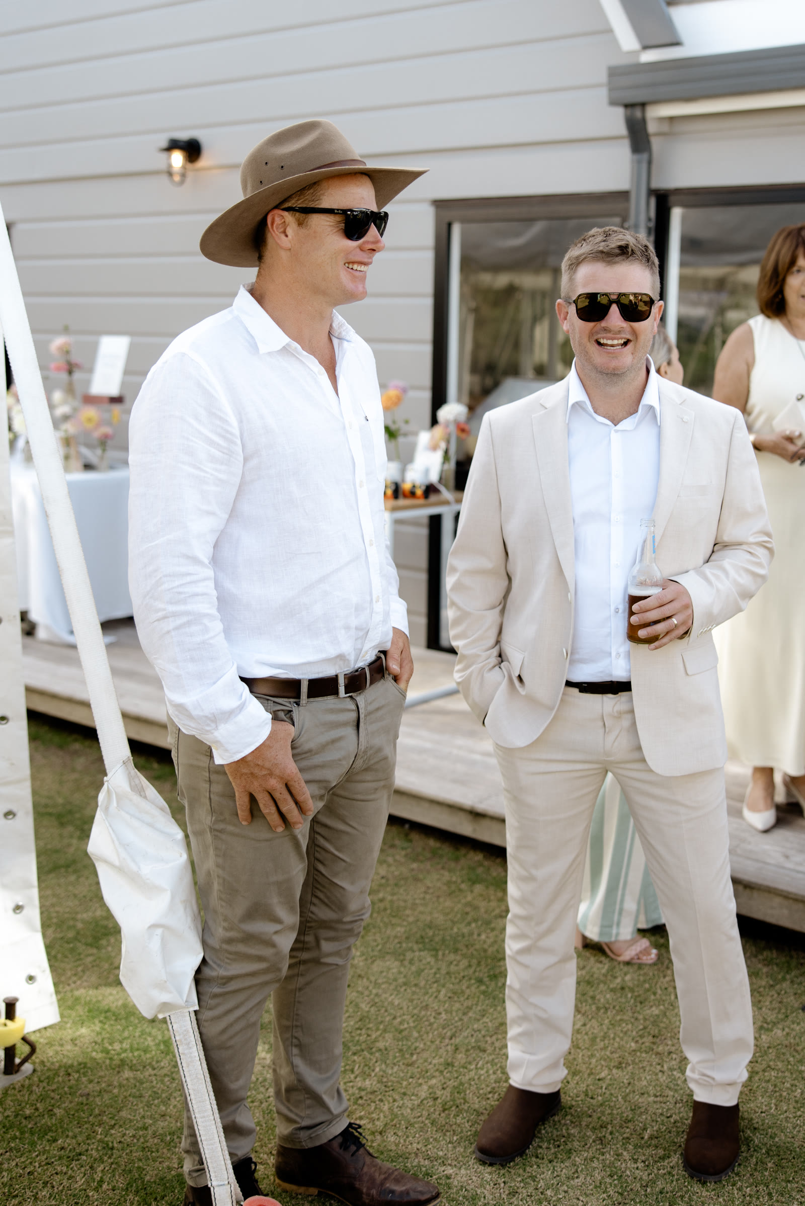 Bryn Sutherland of BMS Construction, in a felt hat and white linen shirt, and Brad Inger of Inger Electrical, in a linen suit with a beer, laughing together on the lawn at Bluestone Estate.