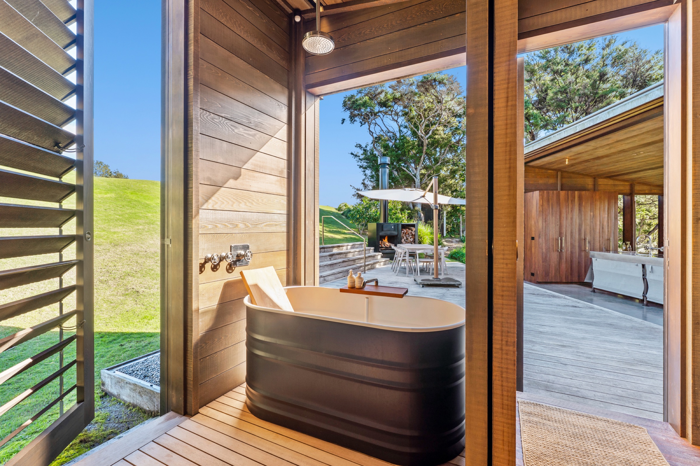 The bath at the Cabins, Bluestone Encampment: a black tub in a cedar-lined nook with a rain shower above, the louvres open to the lawn, and the outdoor fireplace, dining table, and umbrella on the deck between the cabins.