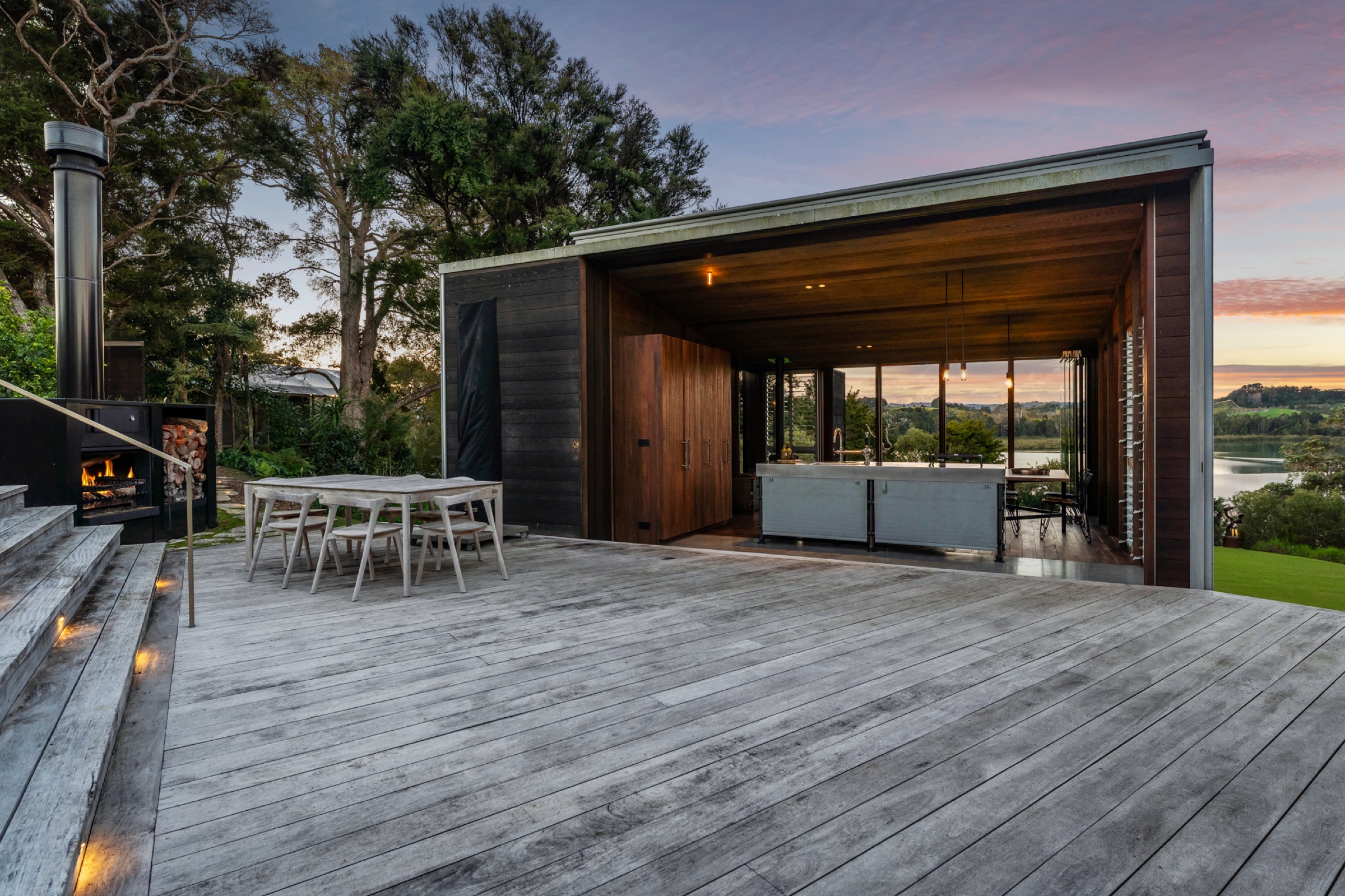 The deck at the Cabins, Bluestone Encampment, at dusk: the outdoor fireplace and dining table, and the living cabin fully open on its long side to the kitchen bench and the inlet beyond.