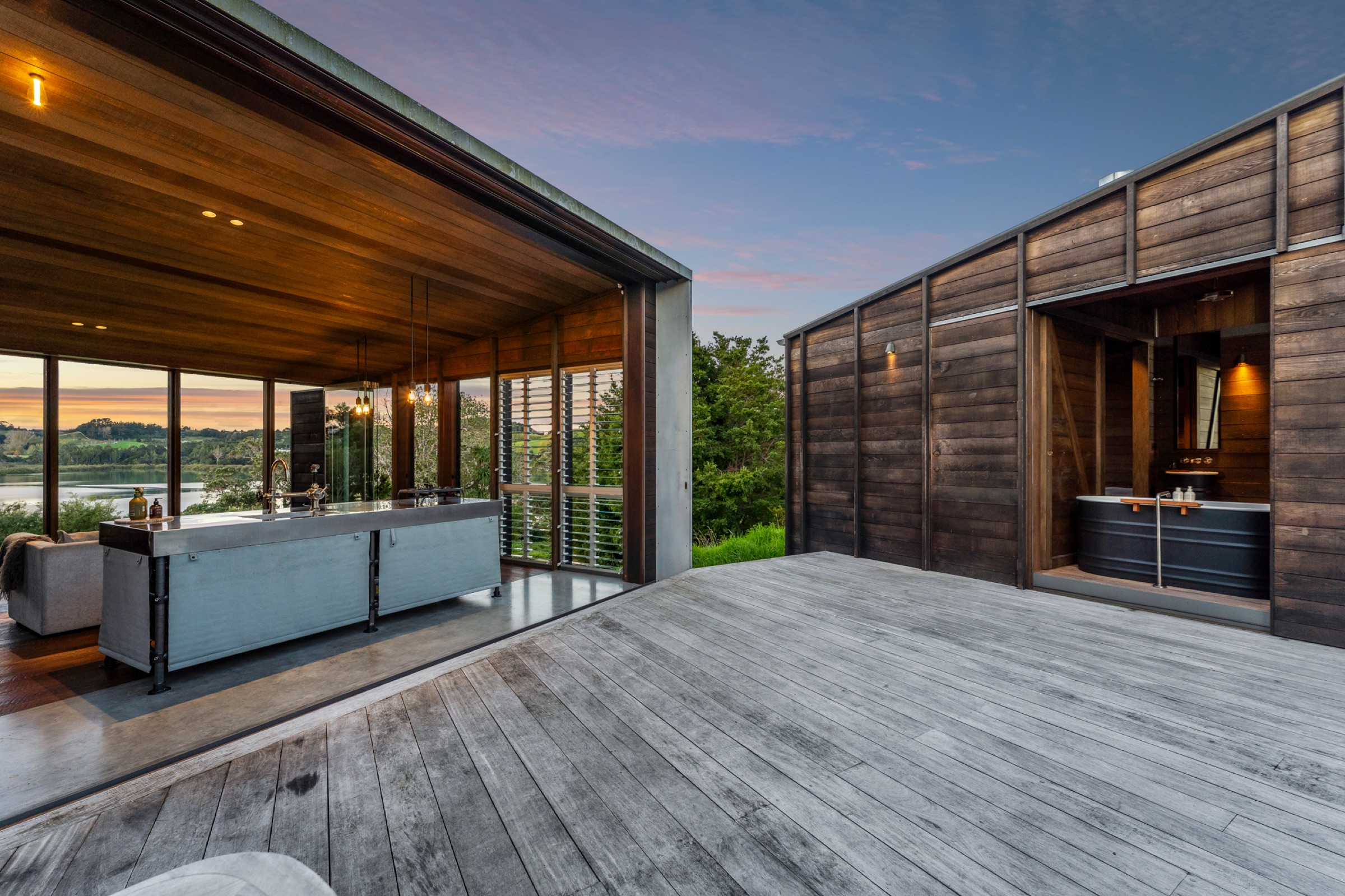 The Cabins at Bluestone Encampment at dusk: the timber deck between the two cedar cabins, the outdoor kitchen bench, and the bath in the open bathroom, with the estuary beyond.