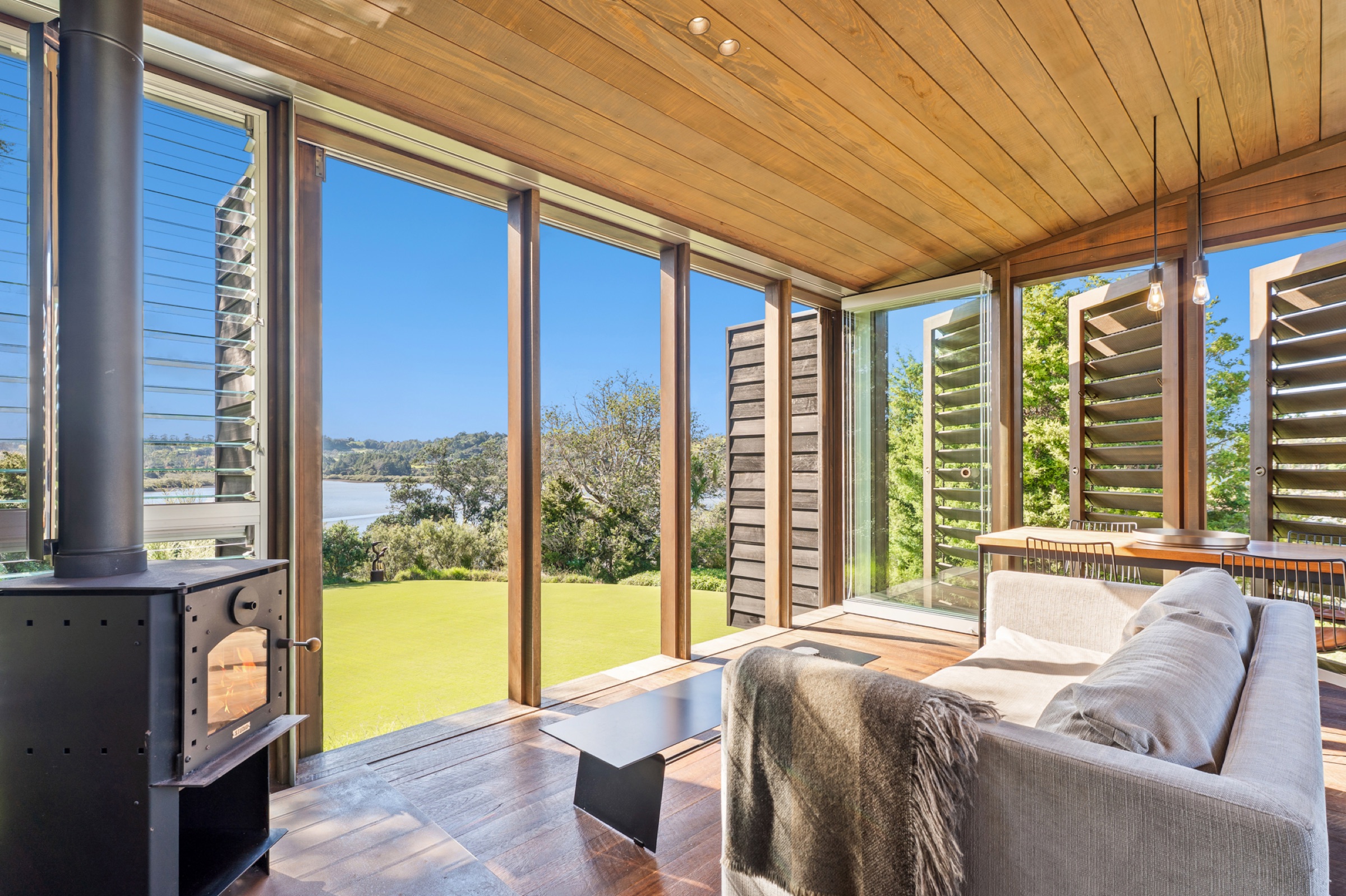 Inside the living cabin of the Cabins at Bluestone Encampment: the wood burner lit, a linen sofa with a wool throw, and the louvres and glass open to the lawn, the bush, and the inlet beyond.