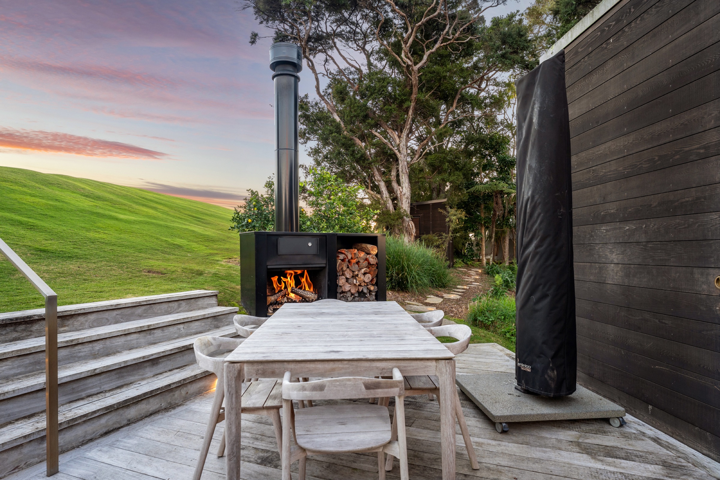 The outdoor fireplace at the Cabins, Bluestone Encampment, lit at sunset beside a stack of firewood, with the weathered timber dining table on the deck, steps up to the lawn, and the bush behind.