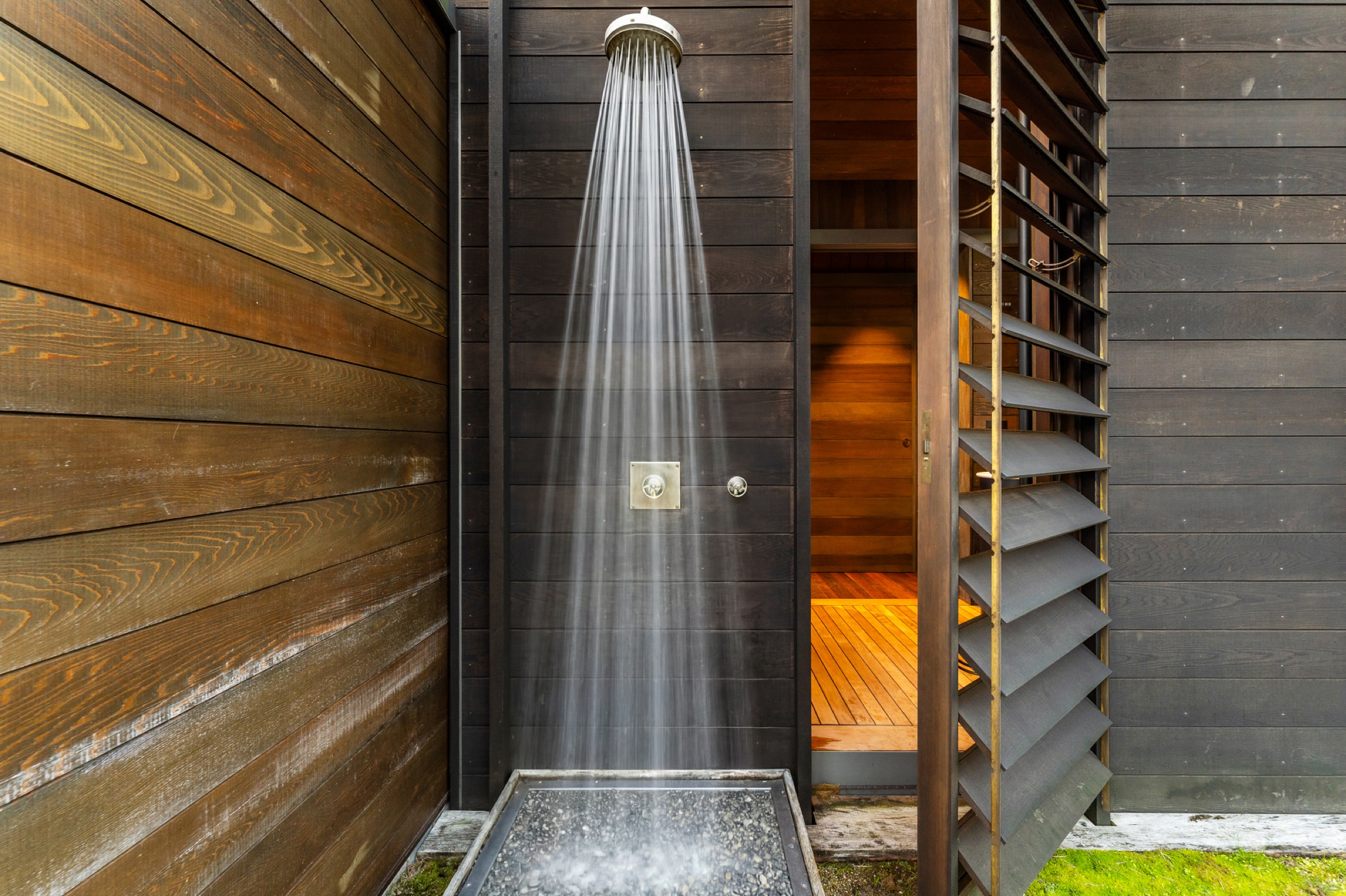 The outdoor shower at the Cabins, Bluestone Encampment: a rain shower running against dark cedar boards over a pebble tray, with the louvred screen open to the timber walkway.