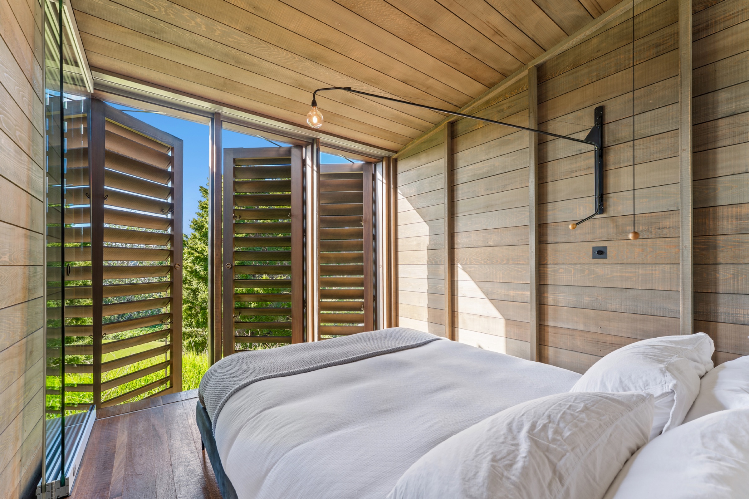 The queen bedroom in the Cabins at Bluestone Encampment: cedar-lined walls and ceiling, white linen and a grey throw, a swing-arm wall lamp, and the timber louvres folded open to the lawn and the bush.