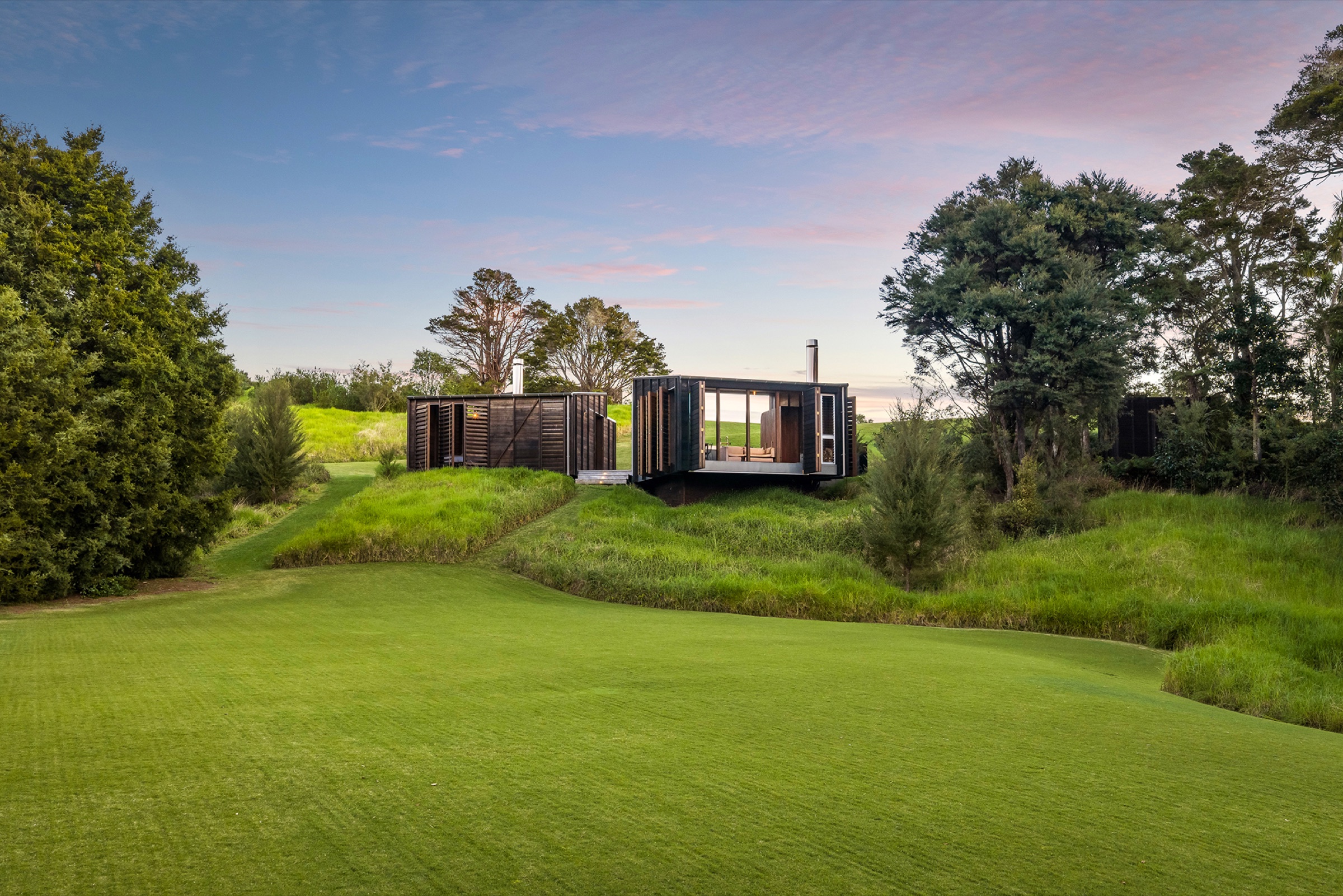 The Cabins at Bluestone Encampment at dusk: two cedar cabins with their louvres open above the lawn, the living cabin lit and glazed on three sides, native bush either side under a pink sky.
