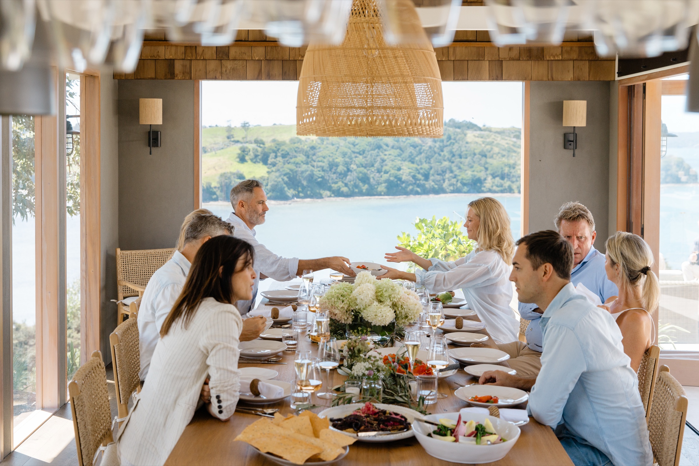 A long lunch at the Cookhouse, Bluestone Ridge: eight guests around the oak table under a rattan pendant, a bowl passed across the hydrangeas, with Kawau Bay and green hills through the open doors.