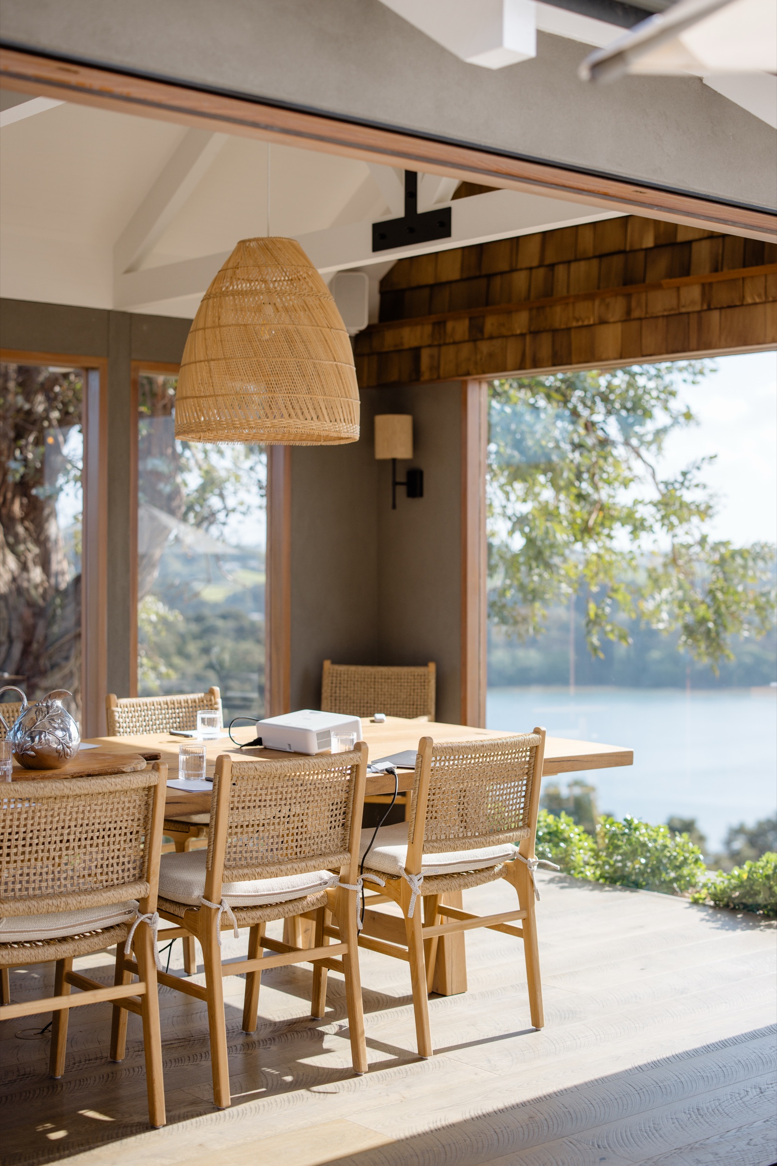 Inside the Cookhouse at Bluestone Ridge: the recycled oak table with its rattan chairs under a woven pendant, cedar shingles above the open doors, and the trees and the water beyond.