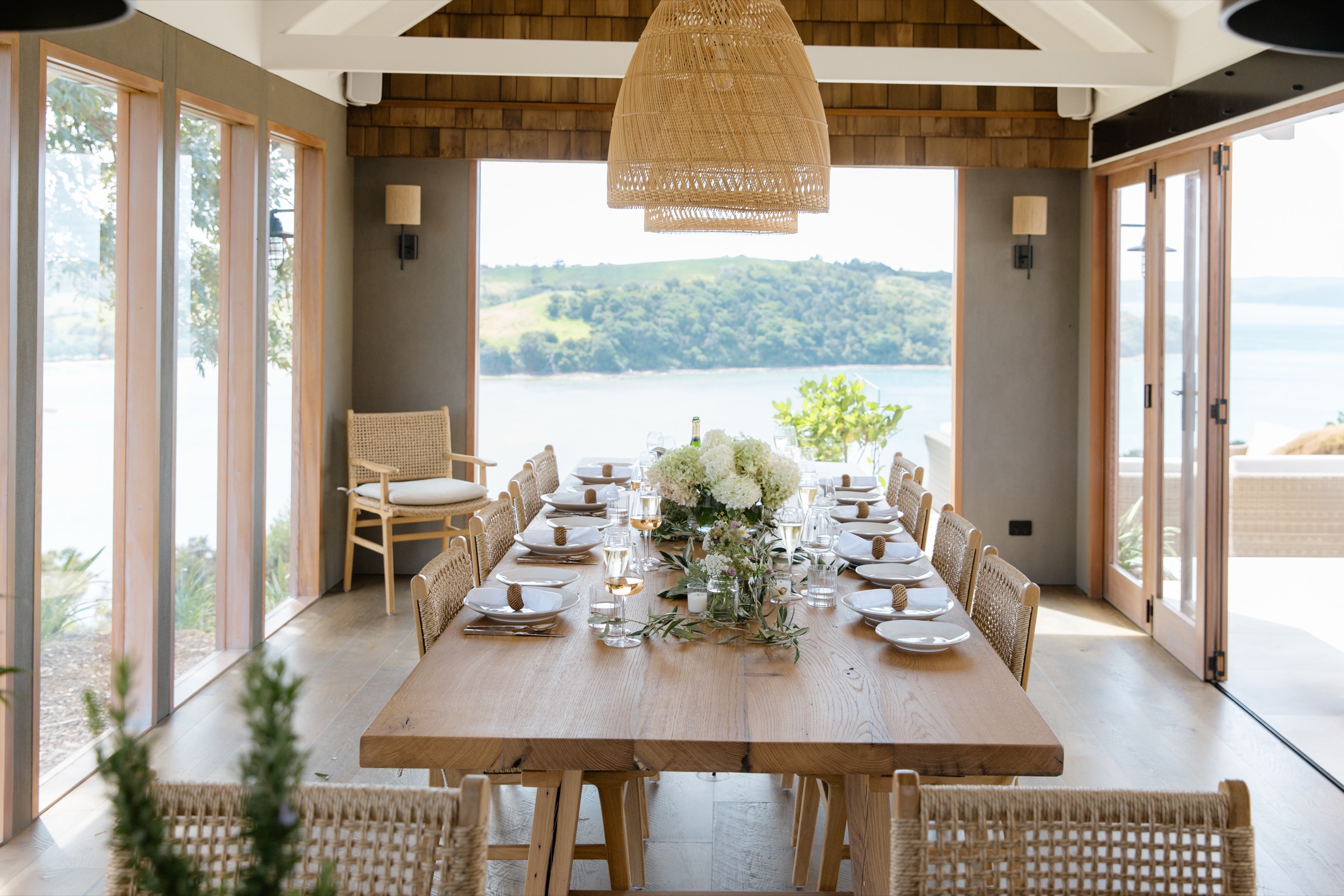 The Cookhouse at Bluestone Ridge: the recycled oak table set for dinner with hydrangeas and olive branches, rattan pendants above, and the doors open to the Hauraki Gulf.