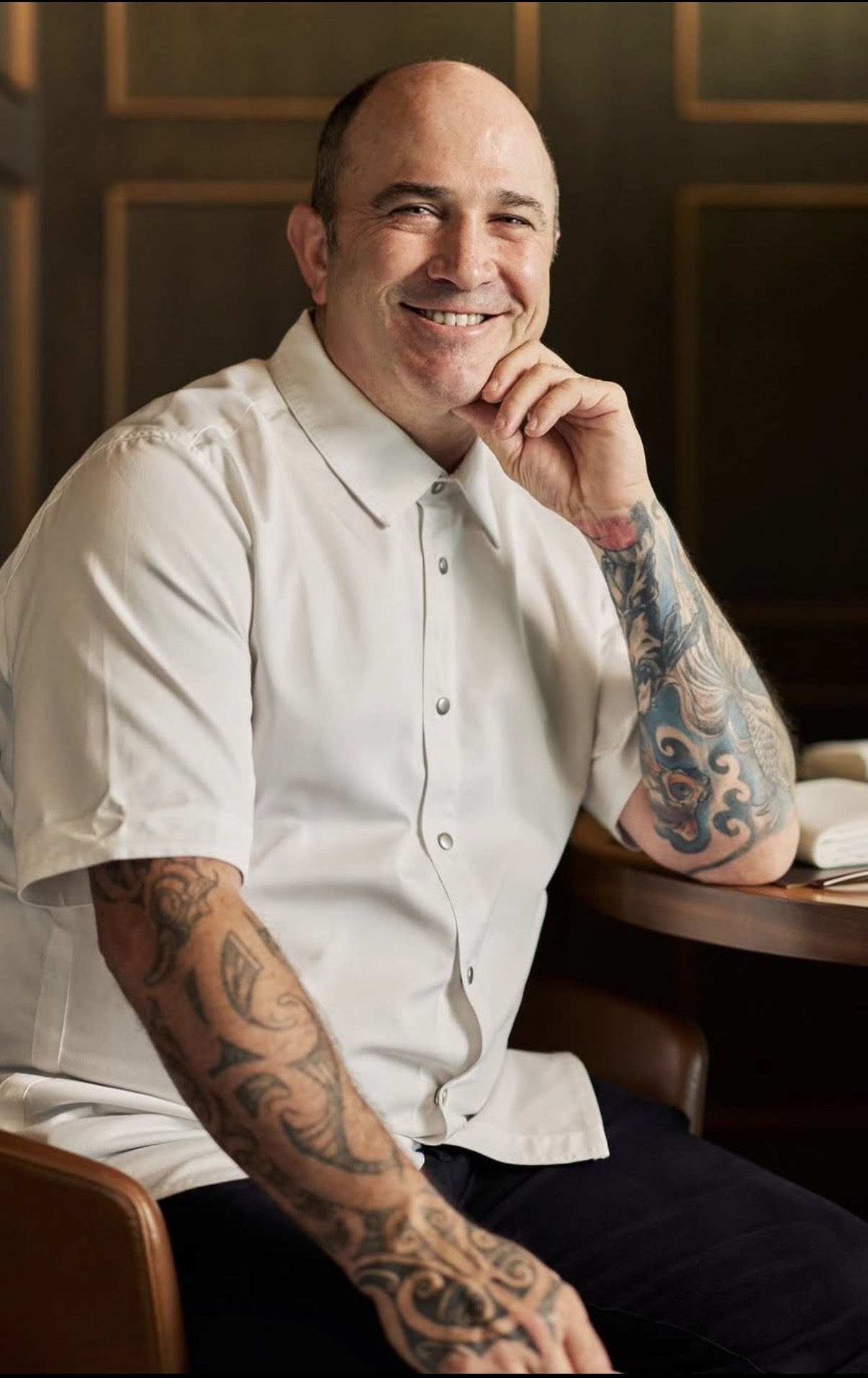 Craig Martin, private chef and founder of Native Kitchen, seated in chef whites with his chin on his hand, smiling, tattooed forearms, dark panelled wall behind.