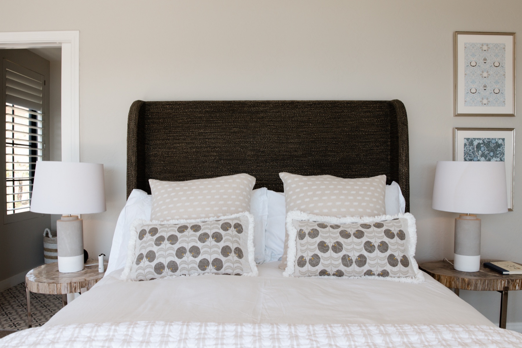 Bedroom 2 at the Farmhouse, Bluestone Ridge: a queen bed with a dark woven headboard, patterned linen cushions, concrete lamps on petrified-wood side tables, framed prints, and shutters at the window.