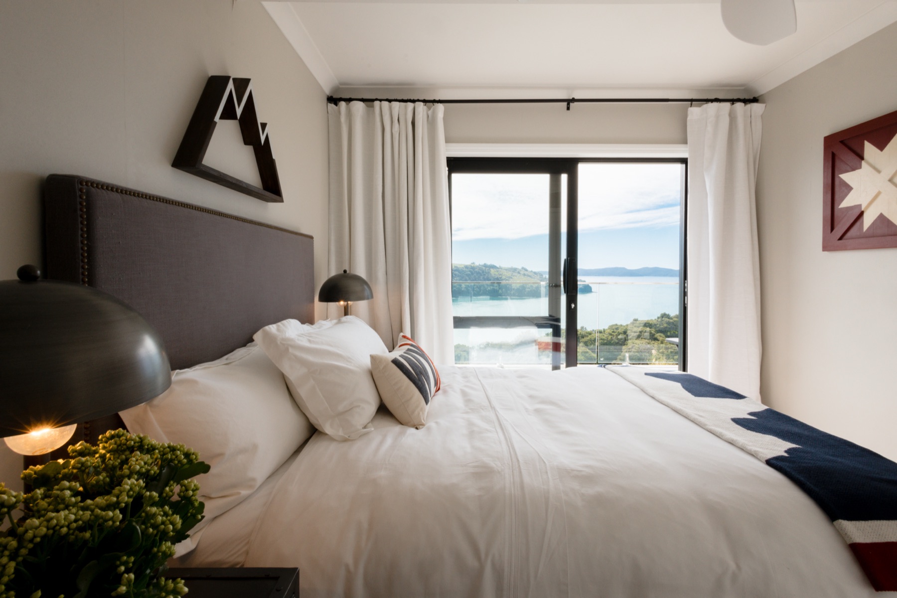 Bedroom 3 at the Farmhouse, Bluestone Ridge: a double bed with a grey studded headboard, a navy throw, black dome lamps, and the sliding door open to the balcony and the Hauraki Gulf.