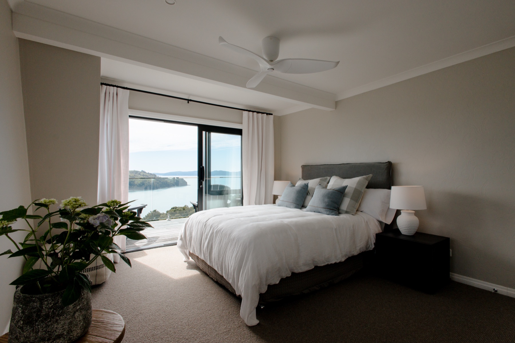 Bedroom 4 at the Farmhouse, Bluestone Ridge: a queen bed with a grey linen headboard and check and blue cushions, a hydrangea in a stone pot, and the sliding doors open to the deck and the Hauraki Gulf.