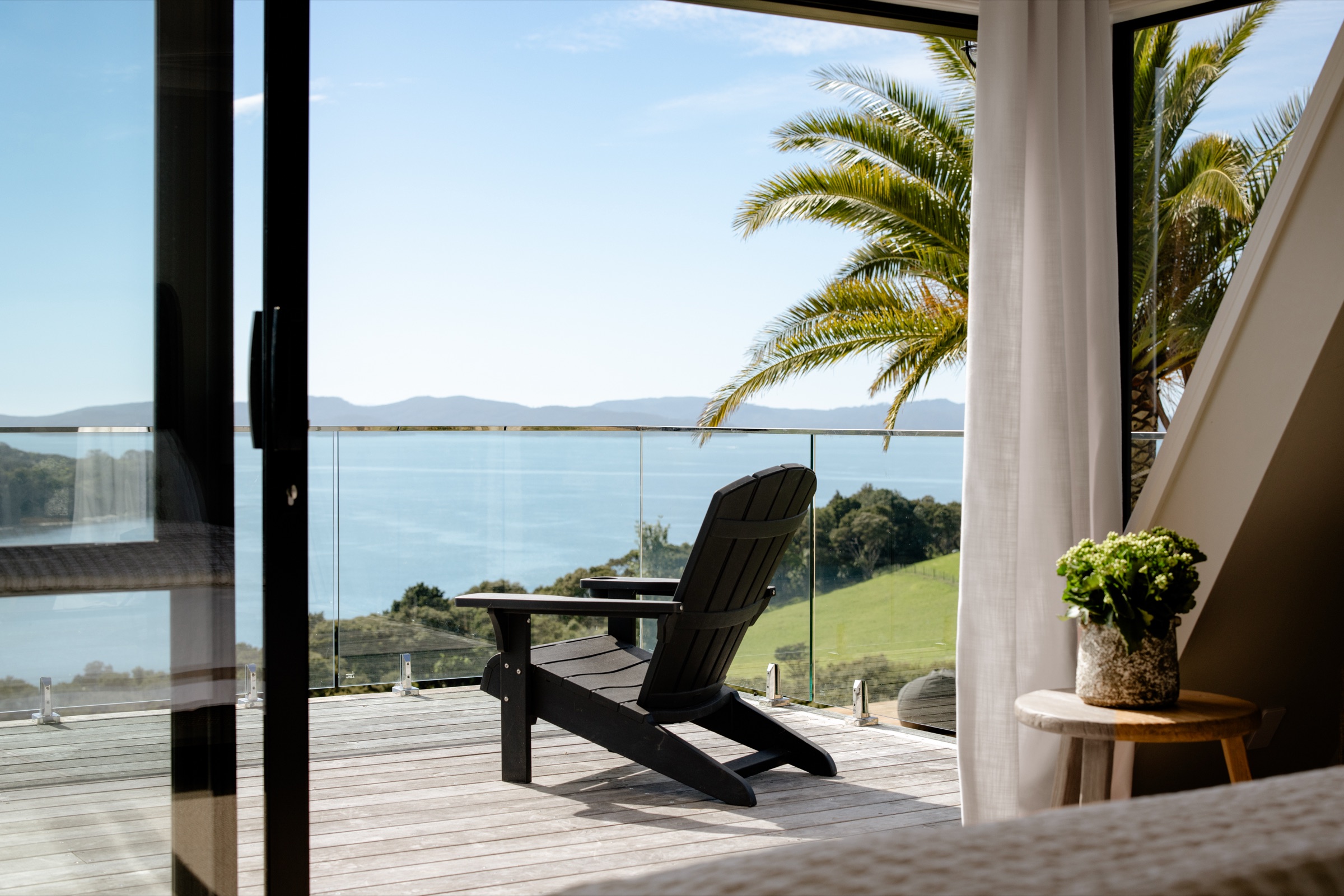 From a bedroom in the Farmhouse at Bluestone Ridge: the sliding door open to the upper terrace, a black Adirondack chair on the deck behind the glass balustrade, a palm beside it, and the Hauraki Gulf and Kawau Island beyond the paddocks.