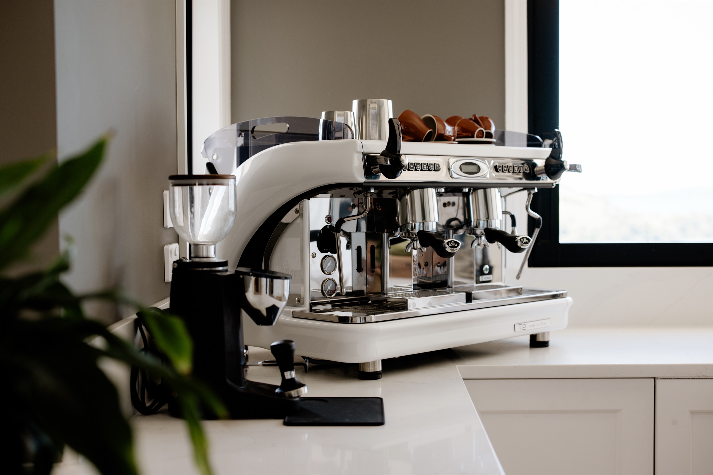 The two-group espresso machine in the Farmhouse kitchen at Bluestone Ridge, with a grinder and tamper beside it and terracotta cups warming on top, by the window.