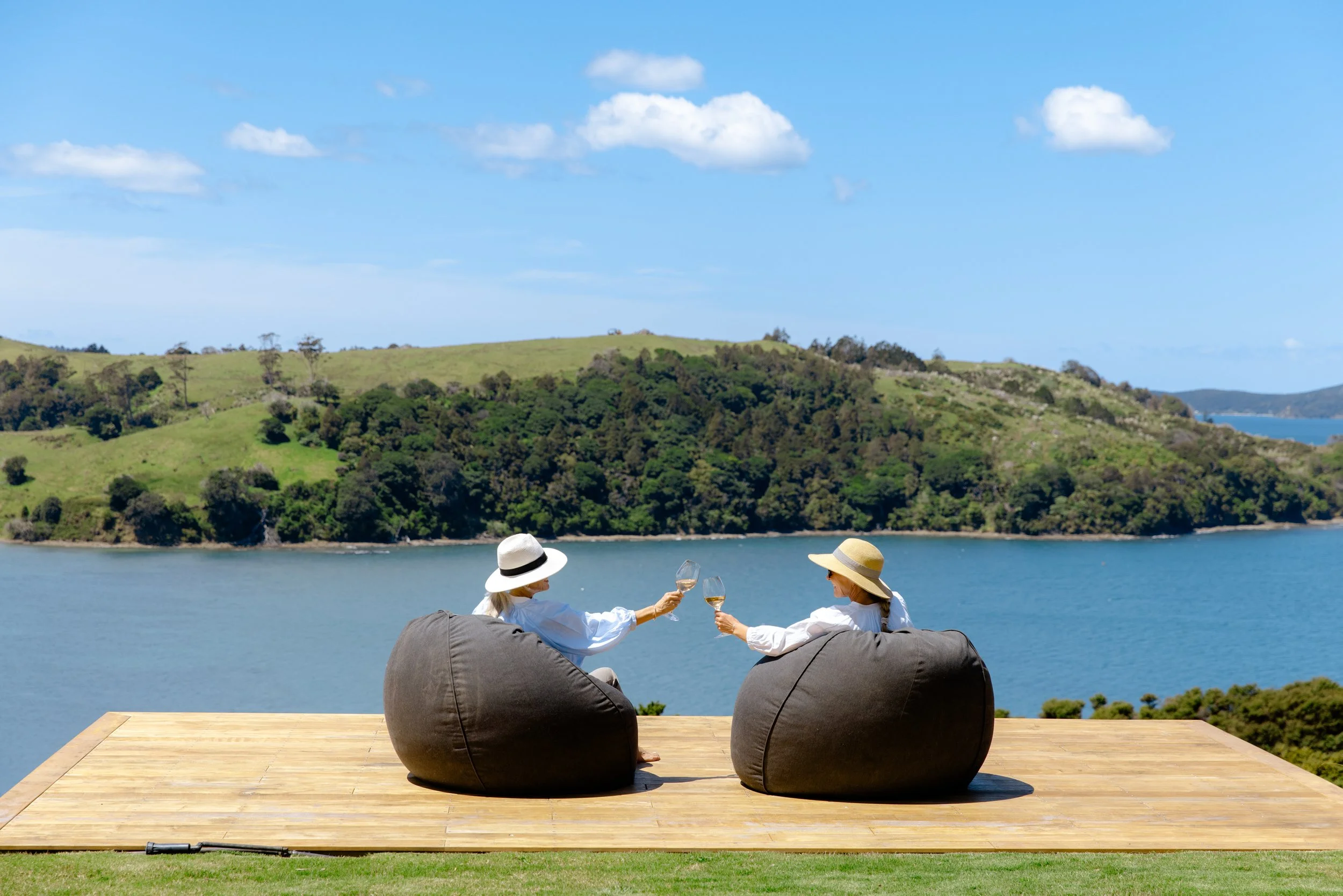 Two guests on beanbags on the Farmhouse deck at Bluestone Ridge, glasses raised to the Hauraki Gulf.