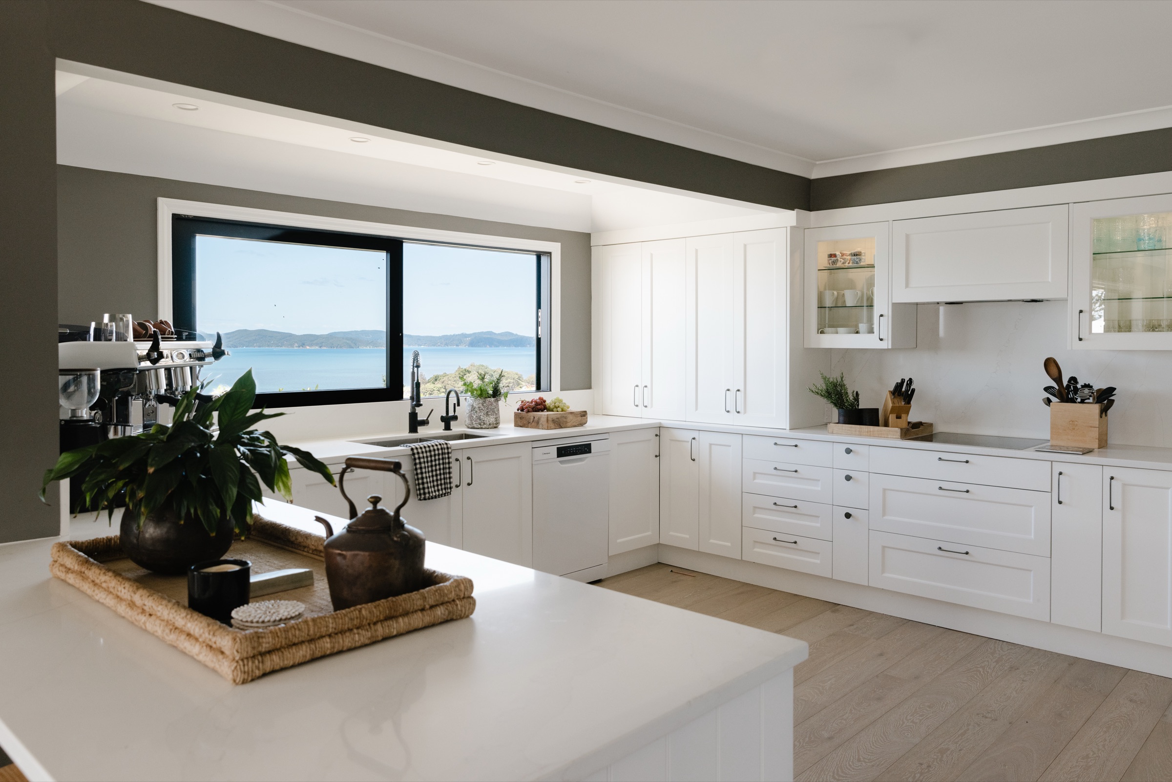 The Farmhouse kitchen at Bluestone Ridge: white cabinetry and stone benches, a copper kettle on a rattan tray on the island, the espresso machine in the corner, and the wide window over the sink looking out to the Hauraki Gulf and Kawau Island.