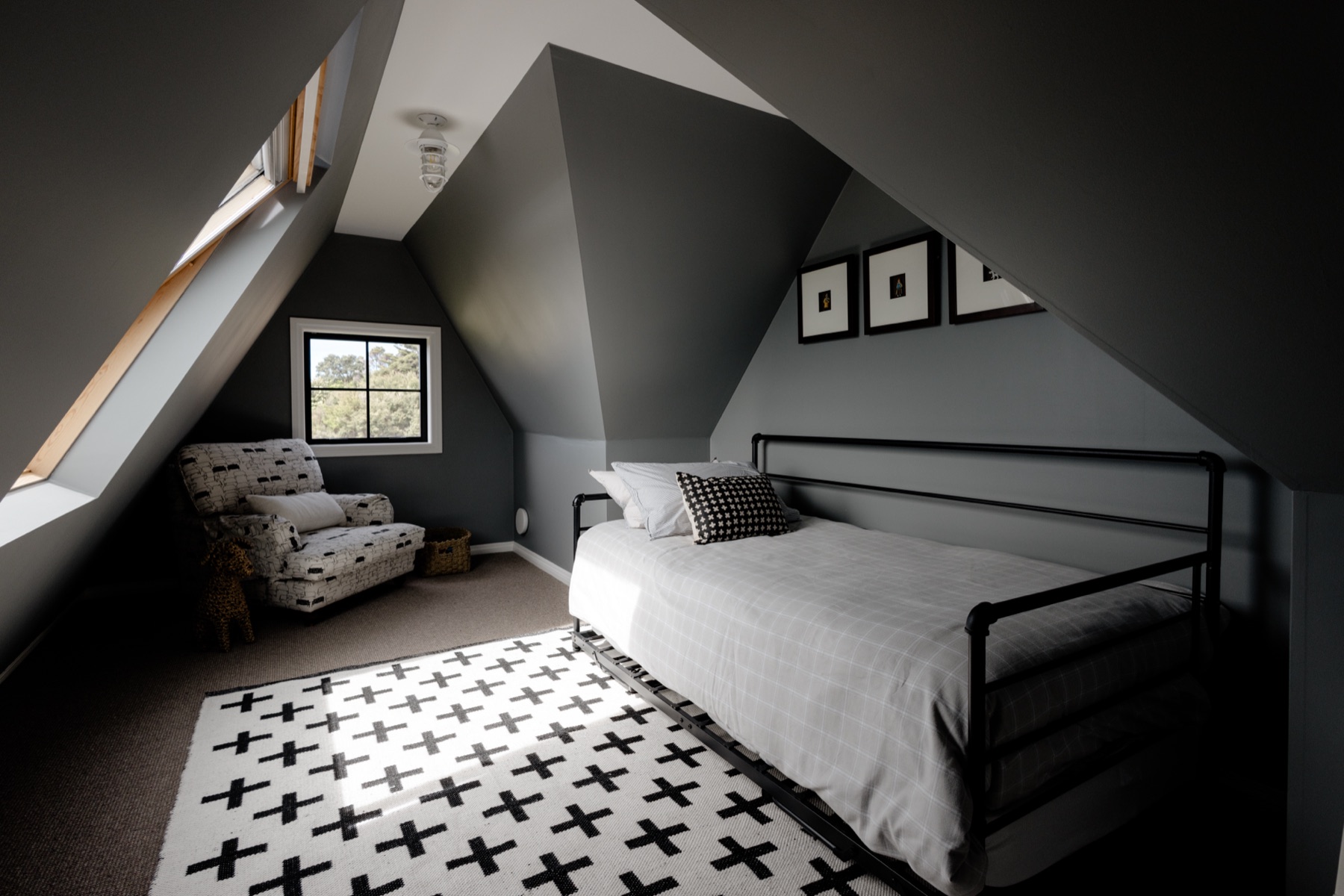 The loft bedroom at the Farmhouse, Bluestone Ridge: a black iron king single with a trundle beneath, under the grey eaves, a cross-patterned rug in a patch of sun from the skylight, a patterned armchair by the window, and three small framed prints.