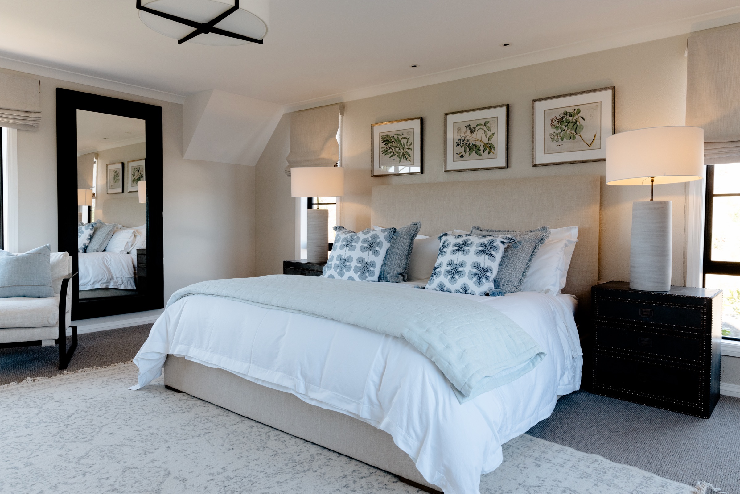 Bedroom 1 at the Farmhouse, Bluestone Ridge: a king bed with a linen headboard, white linen and a pale blue quilt, patterned cushions, three framed botanical prints, ceramic lamps on black bedside chests, and a full-length mirror by the armchair.