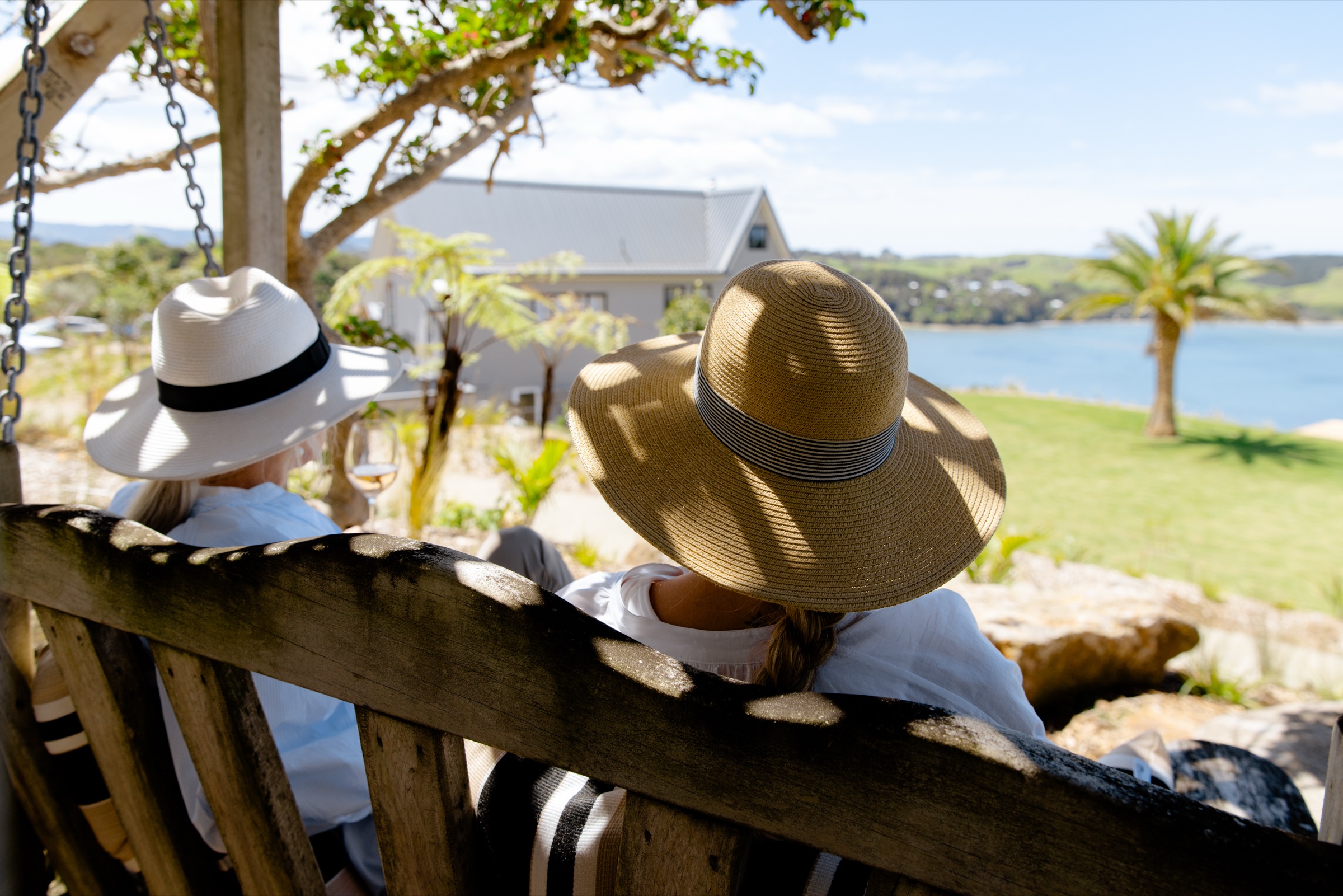 Two guests in sun hats on the timber swing seat under the trees at Bluestone Ridge, a glass of rosé in hand, looking past the Farmhouse and a nīkau to the lawn and the water.
