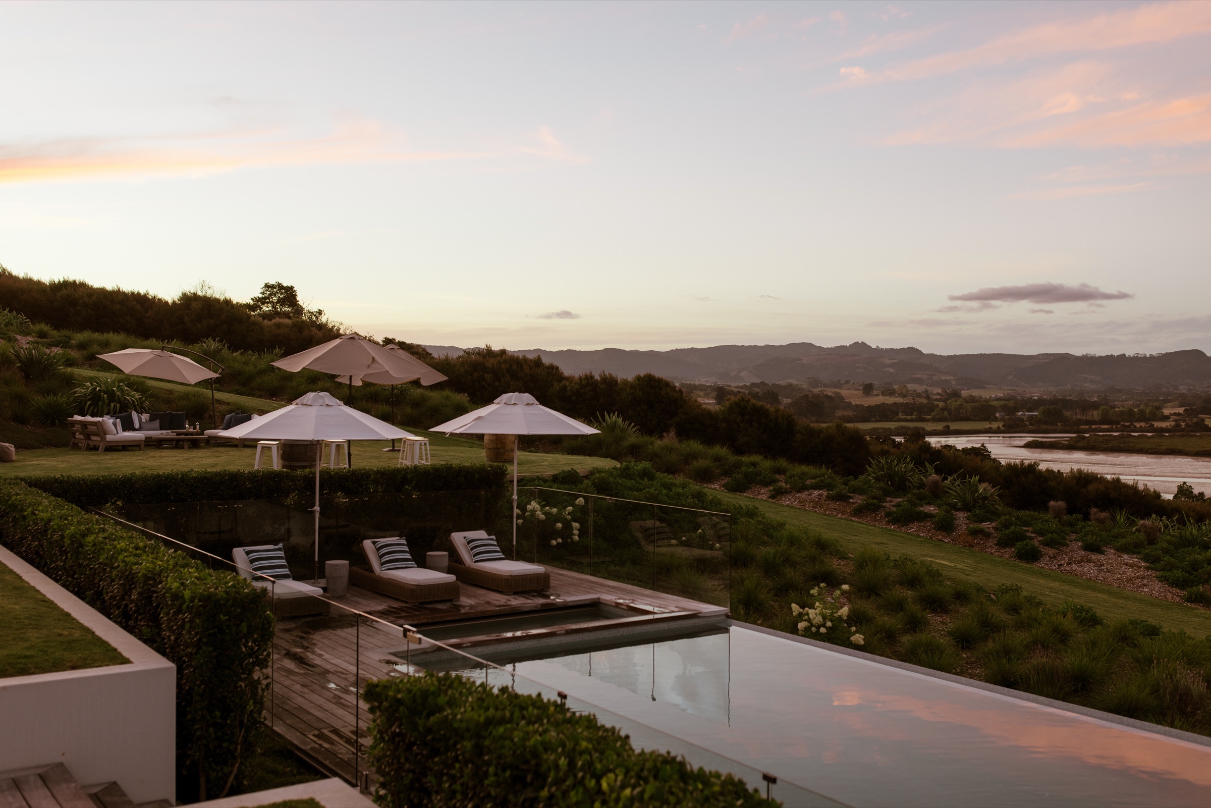 The infinity pool at Bluestone Main Estate at dusk, umbrellas and loungers on the deck, the sky turning pink over the estuary and the hills beyond.