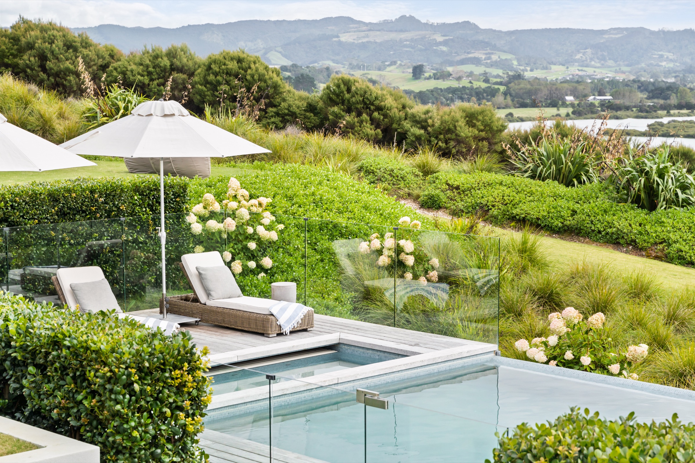 The grounds at Bluestone Main Estate: the pool and spa edged with hydrangeas and native grasses, umbrellas on the lawn, and the estuary and Mt Tamahunga beyond.
