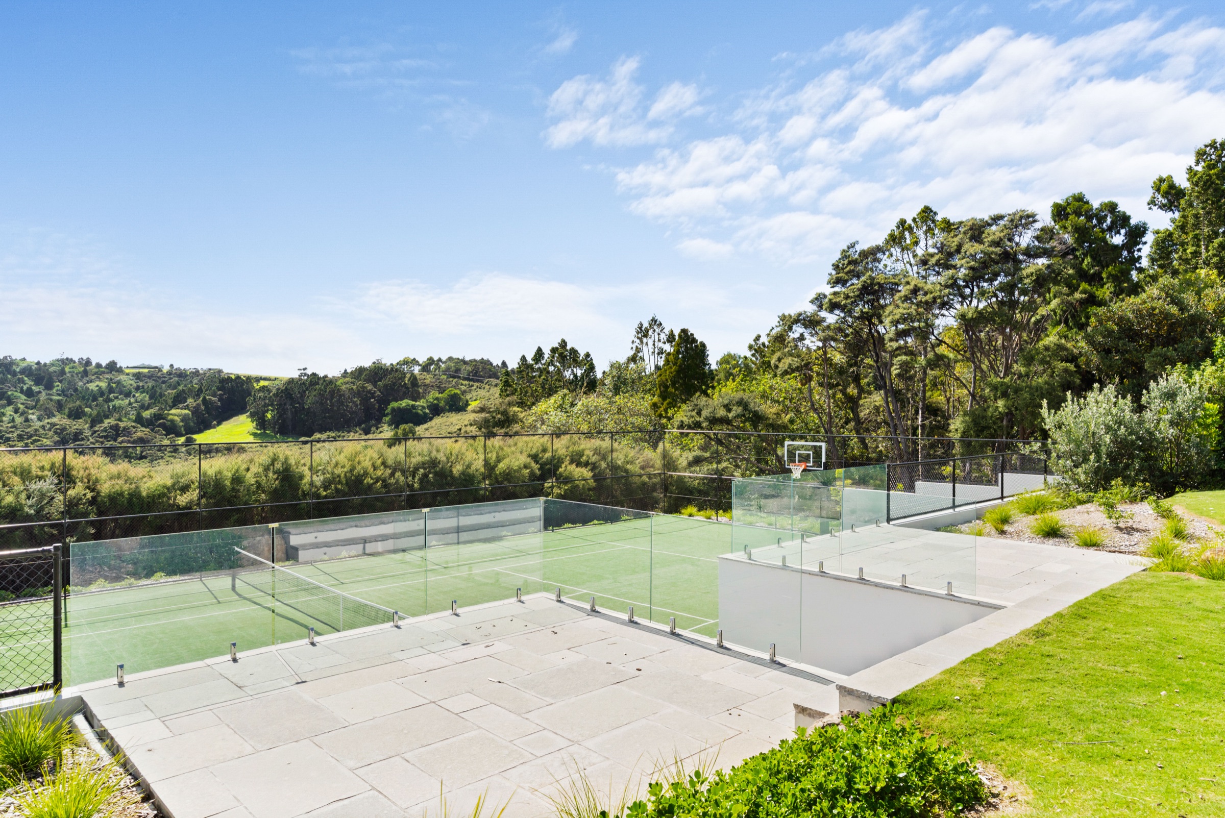 The full-size astroturf tennis court and basketball hoop set into the slope below the Main House at Bluestone Estate, glass-fenced, with native bush and farmland beyond.