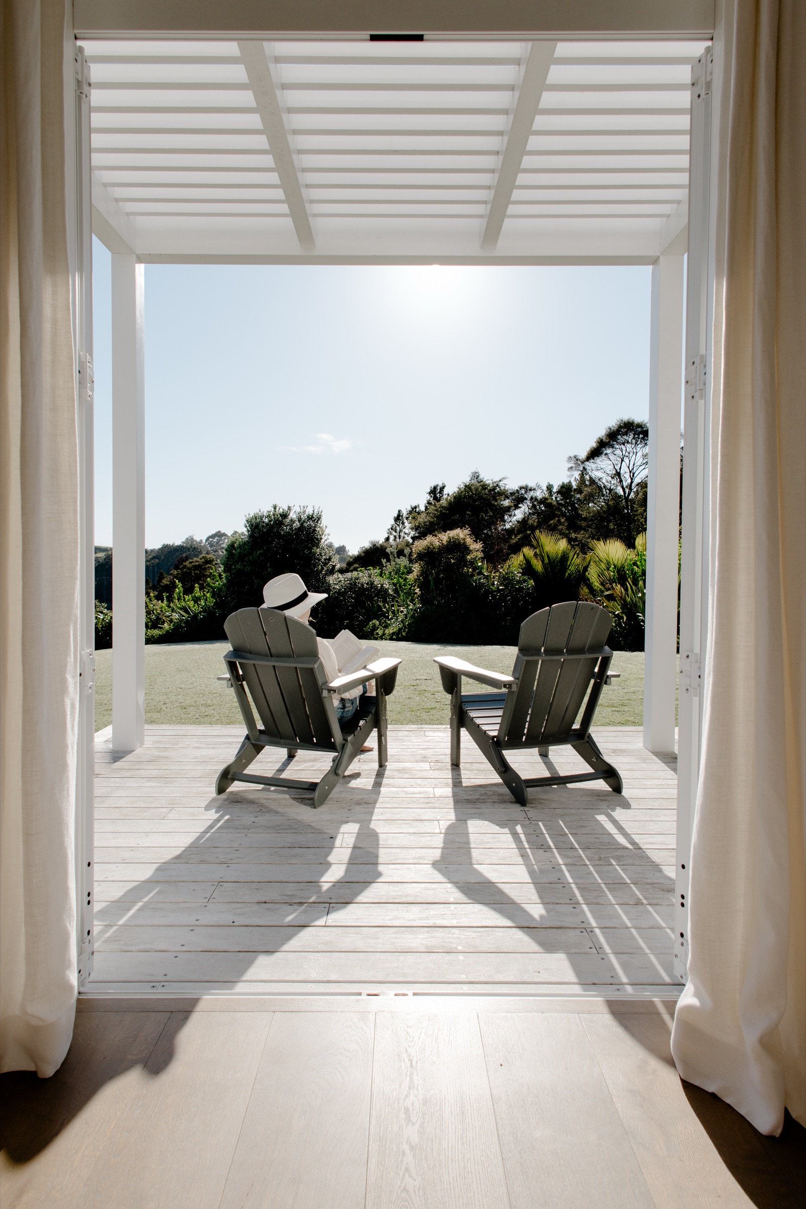 Through the open French doors of a Main House bedroom at Bluestone Estate: a guest in a sun hat reading in an Adirondack chair on the deck, the lawn and native bush in bright sun.