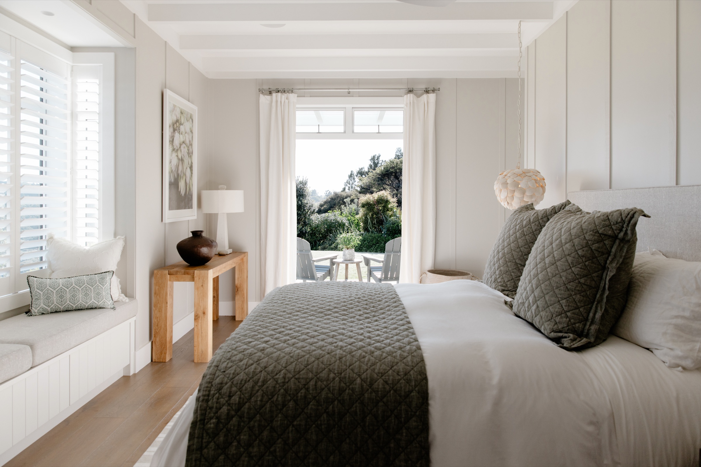 A ground-floor bedroom in the Main House at Bluestone Estate: a window seat with shutters, a quilted throw on the bed, a shell pendant light, and French doors open to the deck, the Adirondack chairs, and the bush.