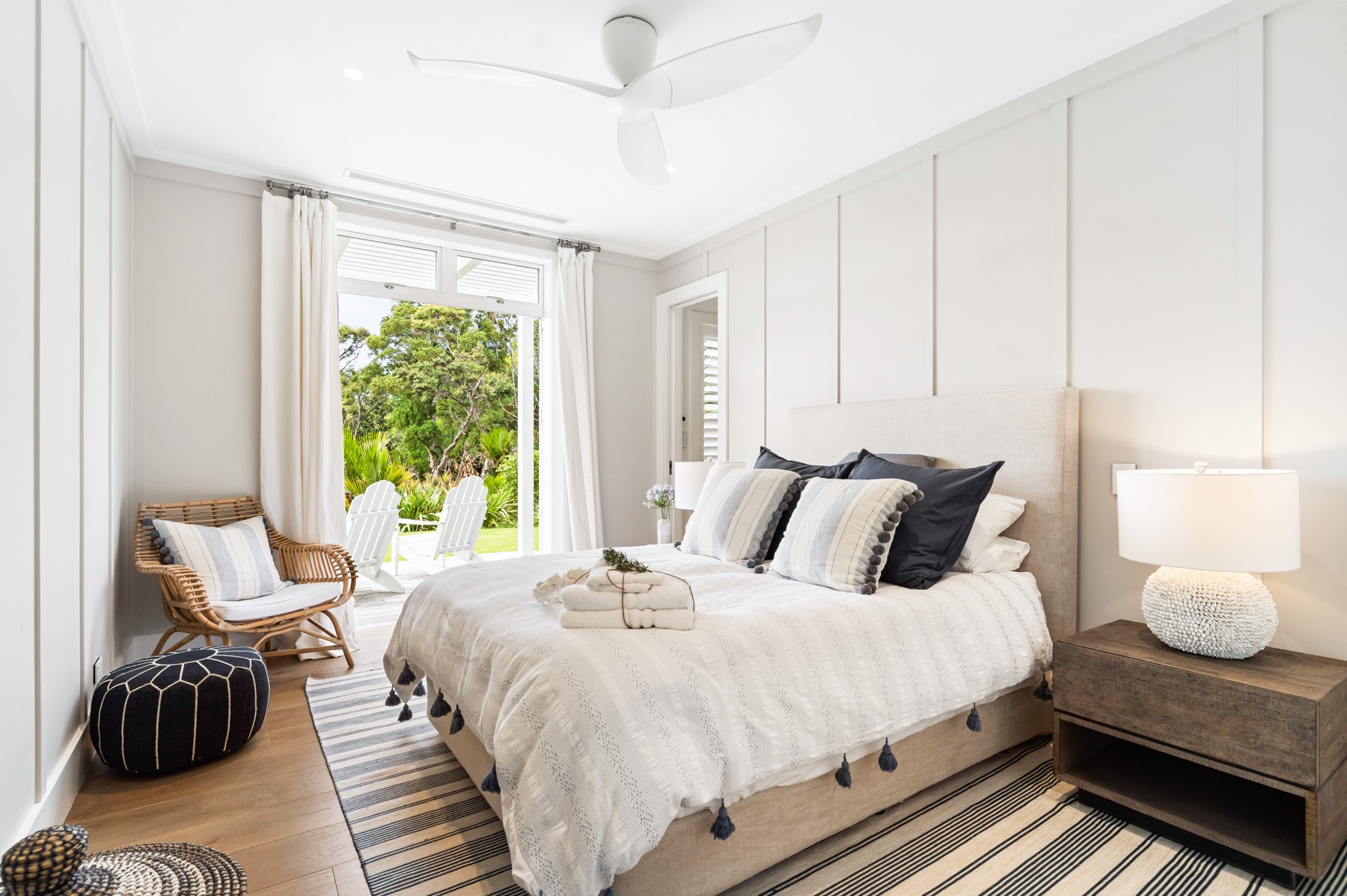 The Blue Bedroom at the Main House, Bluestone Estate: a queen bed with navy and striped cushions and a tasselled throw, a shell lamp, a rattan chair and a navy pouf, and French doors open to the deck and two white Adirondack chairs.