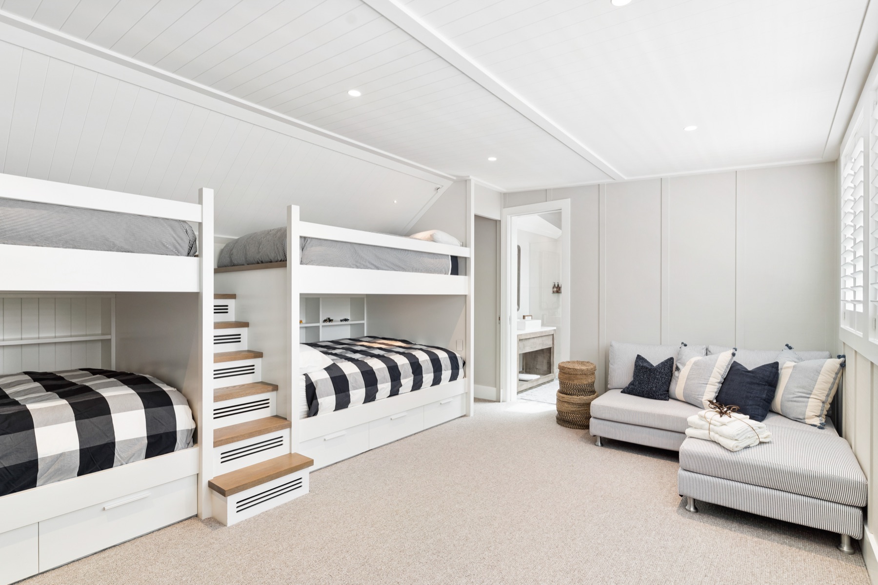 The loft bunk room at the Main House, Bluestone Estate: two built-in queen bunks with black and white check covers and stairs with drawers between them, a striped sofa under the shuttered window, and the ensuite through the door.