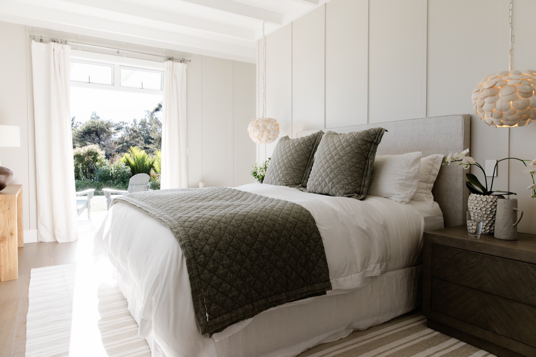 The Primary Bedroom at the Main House, Bluestone Estate: a king bed with a linen headboard, olive quilted throw and cushions, shell pendants either side, board-and-batten walls, and French doors open to the garden and the Adirondack chairs outside.