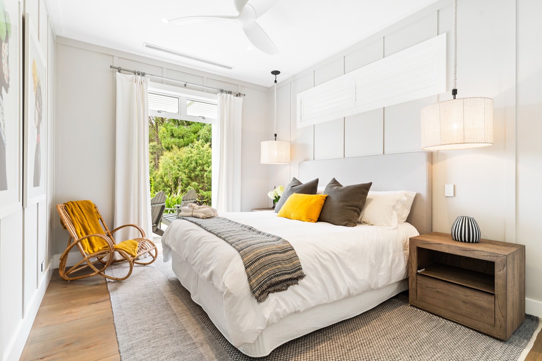 The Yellow Bedroom at the Main House, Bluestone Estate: a king bed with a yellow cushion and a striped throw, linen pendants either side, a rattan rocking chair with a mustard blanket, and French doors open to the garden.