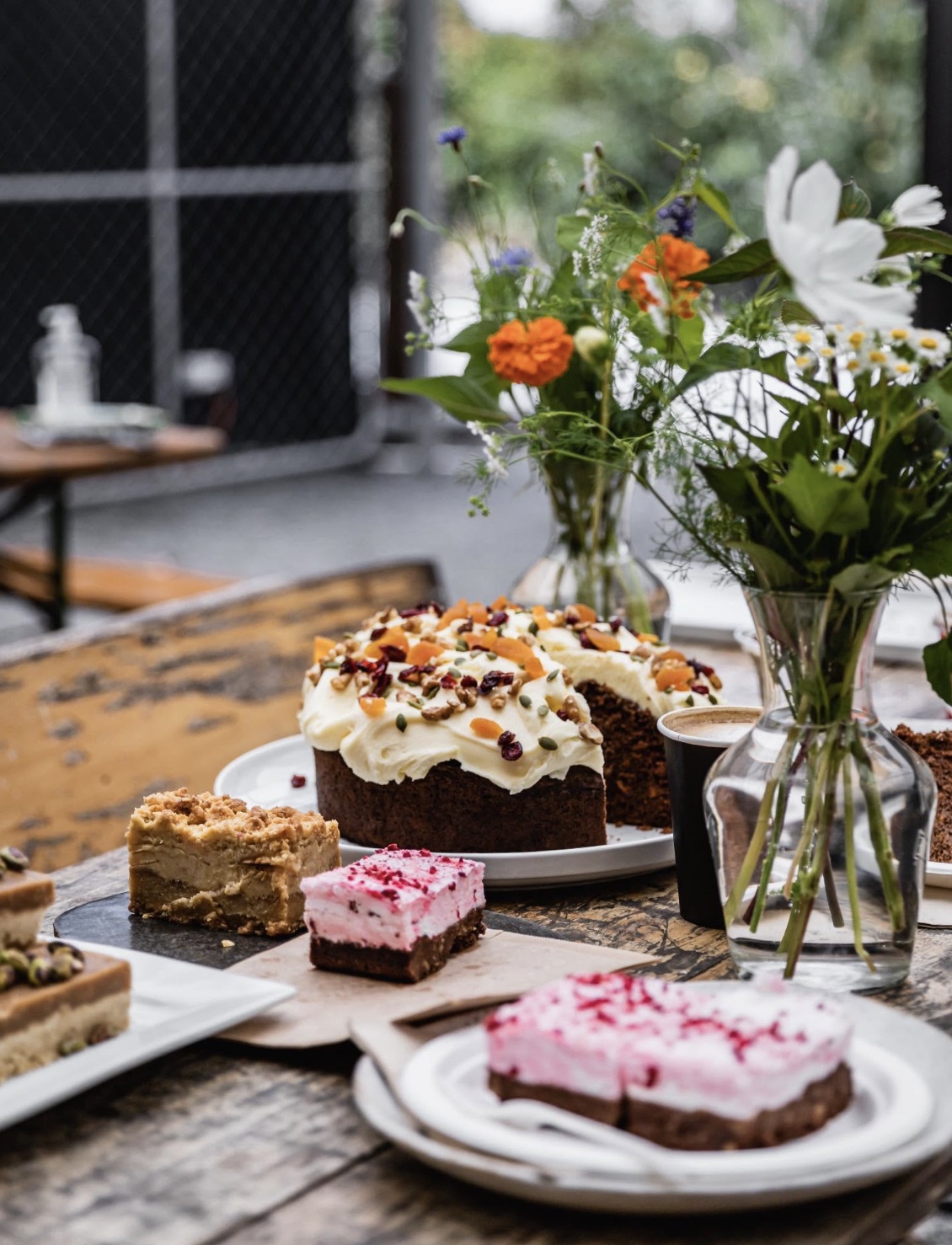 Ravish, Matakana: a carrot cake topped with dried apricot, walnuts, and seeds, pink-iced slices, and a coffee on a worn timber table with jars of wildflowers.