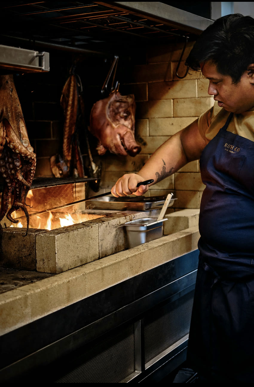 Rothko at Sculptureum, Matakana: a chef in a navy apron tending the wood-fired grill, with octopus, fish, and a pork head hanging to smoke above the flames.