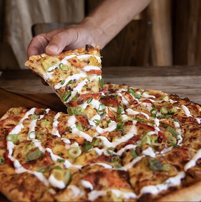 Rusty Pelican, Matakana: a hand lifting a slice from a pizza topped with spring onion, roast pepper, and drizzled aioli, on a timber board.