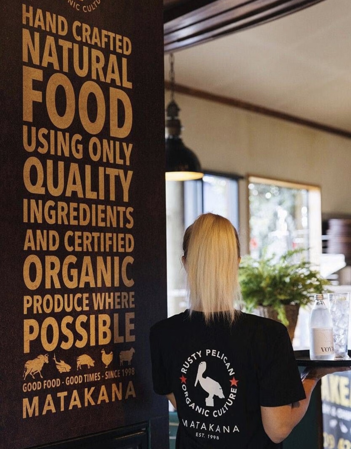 Rusty Pelican, Matakana: a waiter carrying a tray past the painted wall that reads hand crafted natural food using only quality ingredients and certified organic produce where possible.